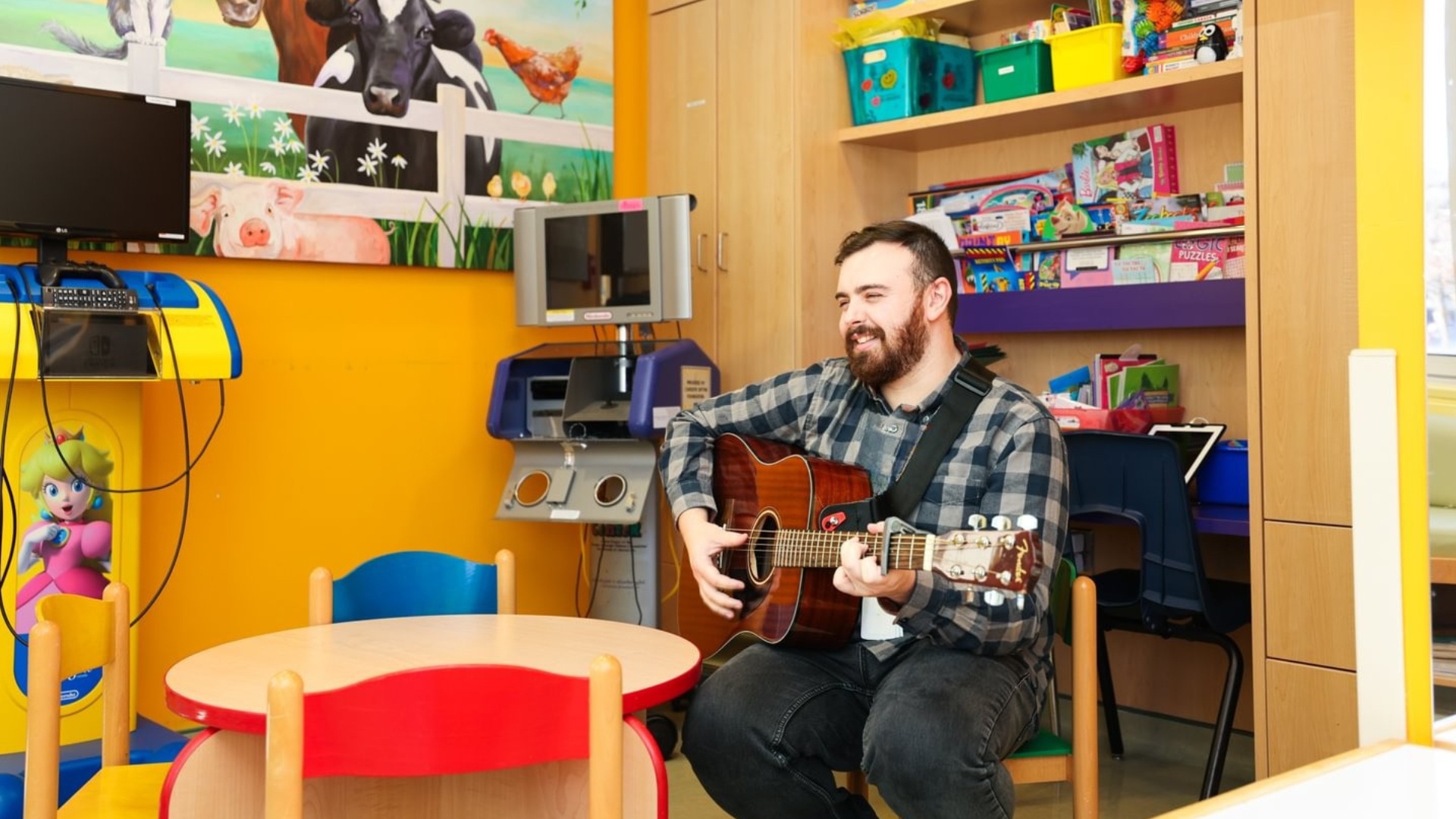 A man sits in a colorful playroom, playing an acoustic guitar and smiling, surrounded by children's toys and books.