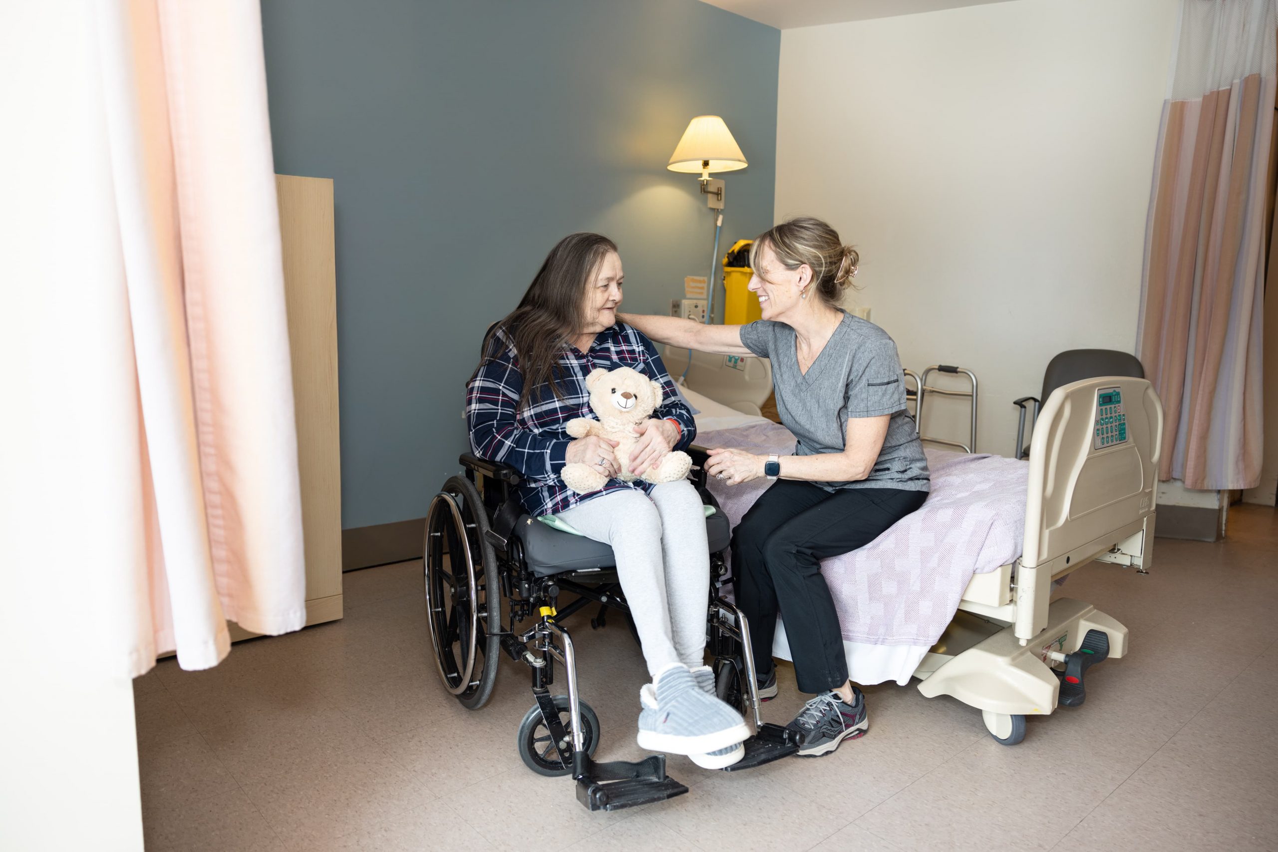 A caregiver sits beside a woman in a wheelchair holding a teddy bear in a hospital room.