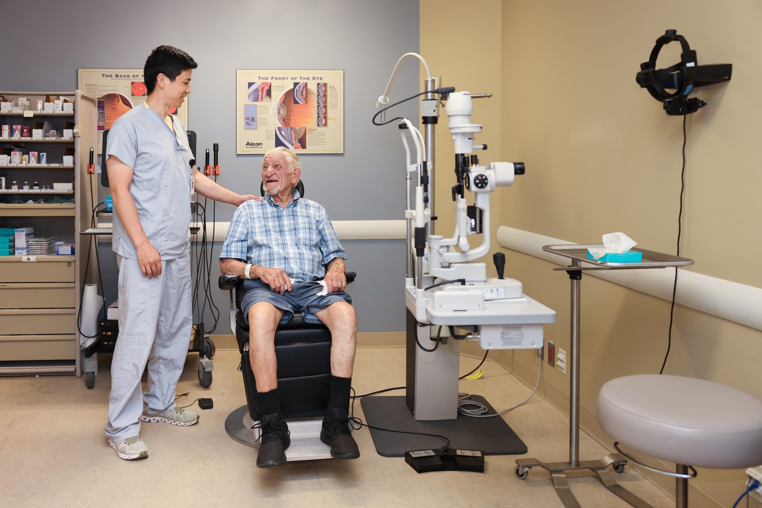 An elderly man sits in an eye exam room, smiling at a healthcare professional standing beside him.