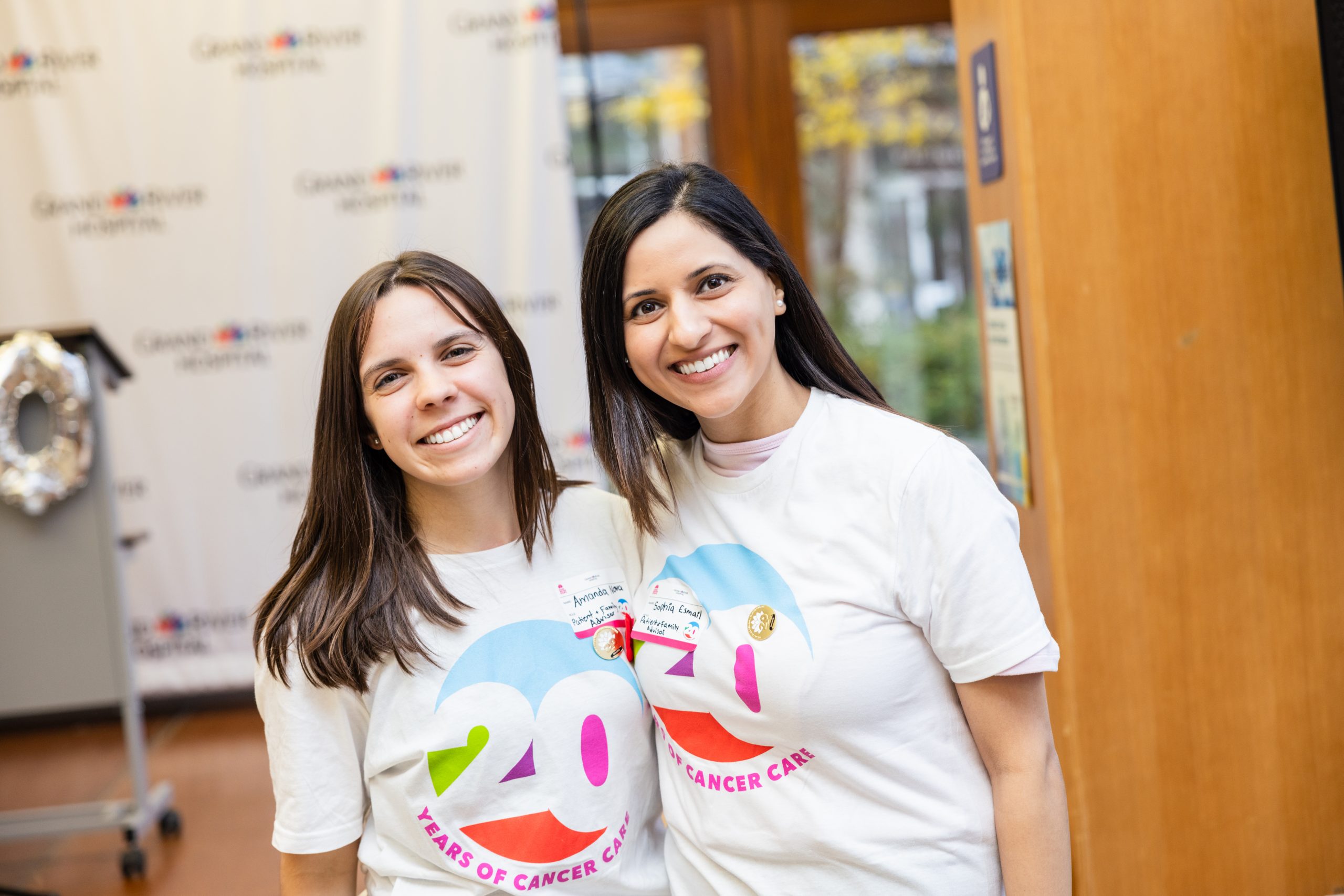 Two women smiling indoors, wearing white shirts with a colorful "20 Years of Cancer Care" logo.