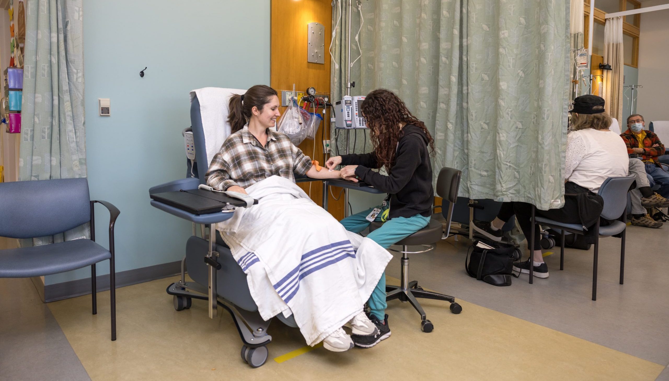 A nurse checks a woman’s arm while she sits in a medical chair covered with a blanket in a clinic room.