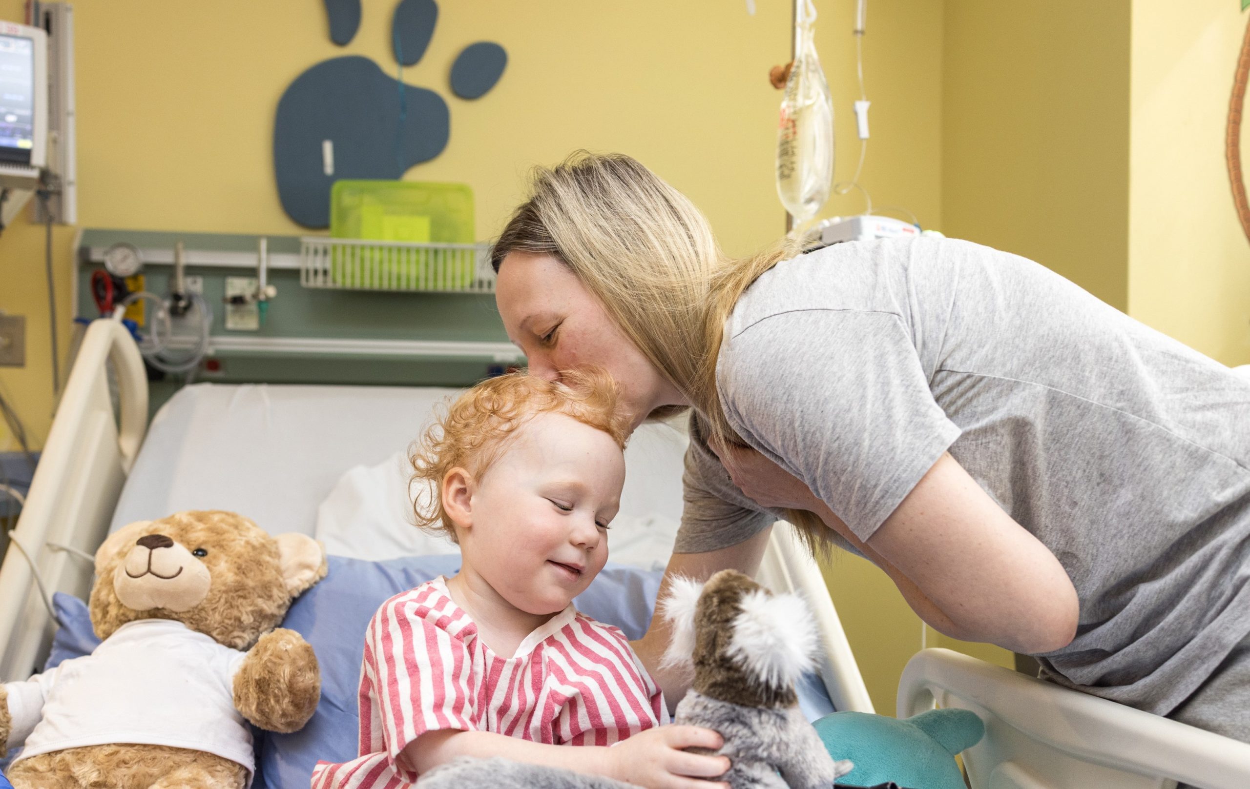 An adult kisses a smiling child on the head as the child sits in a hospital bed with stuffed animals.