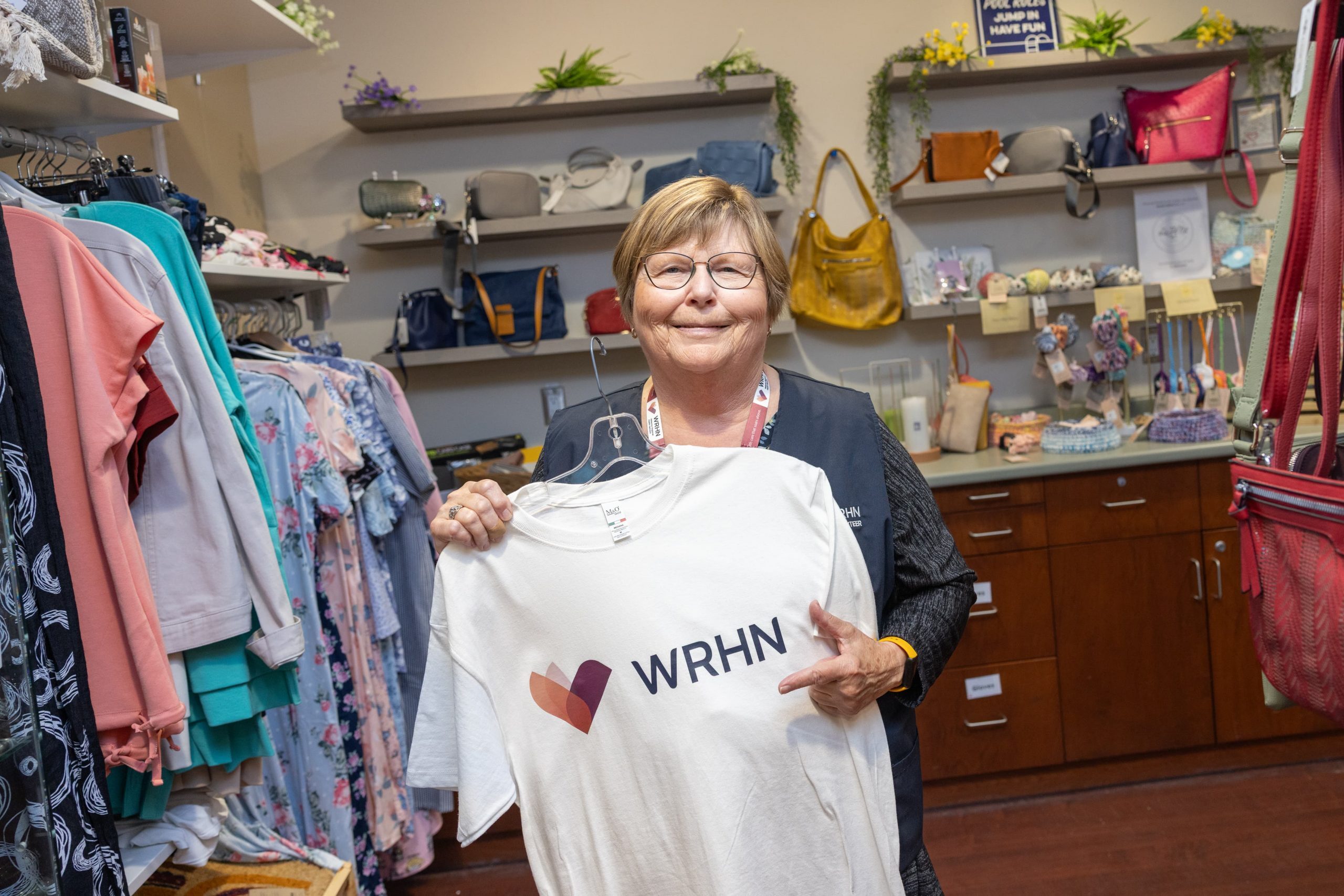Woman holding a white WRHN t-shirt in a store with clothing, handbags, and shelves in the background.