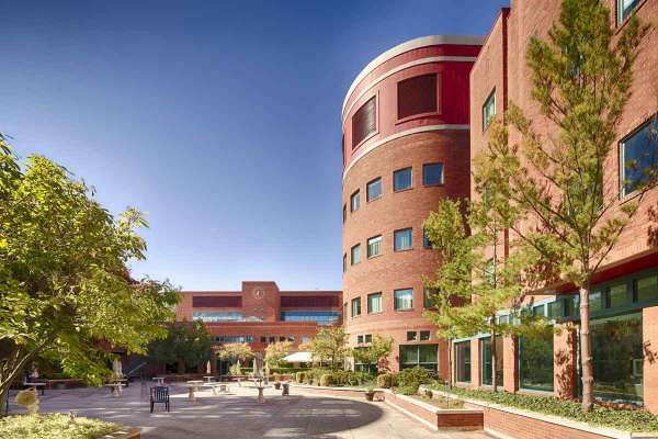 Red brick academic building with rounded and rectangular sections, trees, and a central open courtyard under a clear sky.