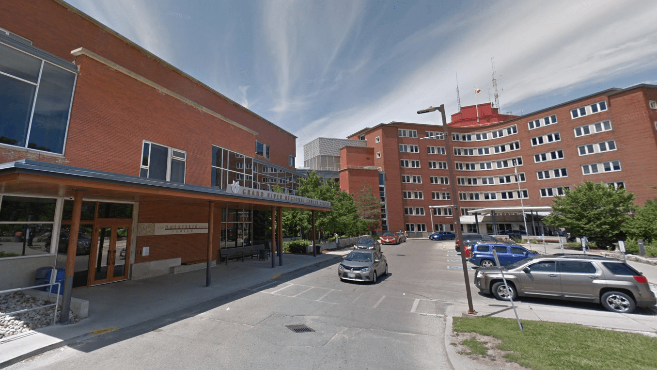 A brick hospital building with an entrance canopy, parked cars, and a partly cloudy sky overhead.