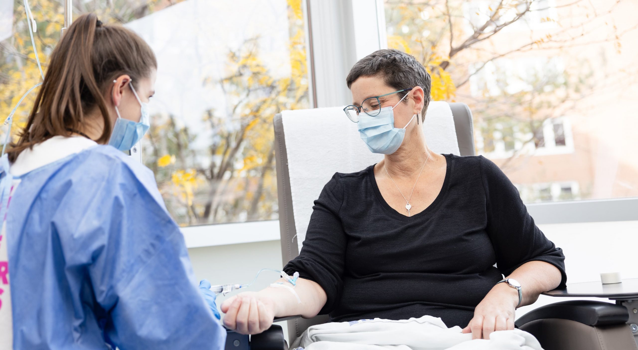 A healthcare worker administers an IV to a seated patient wearing a mask in a medical setting.