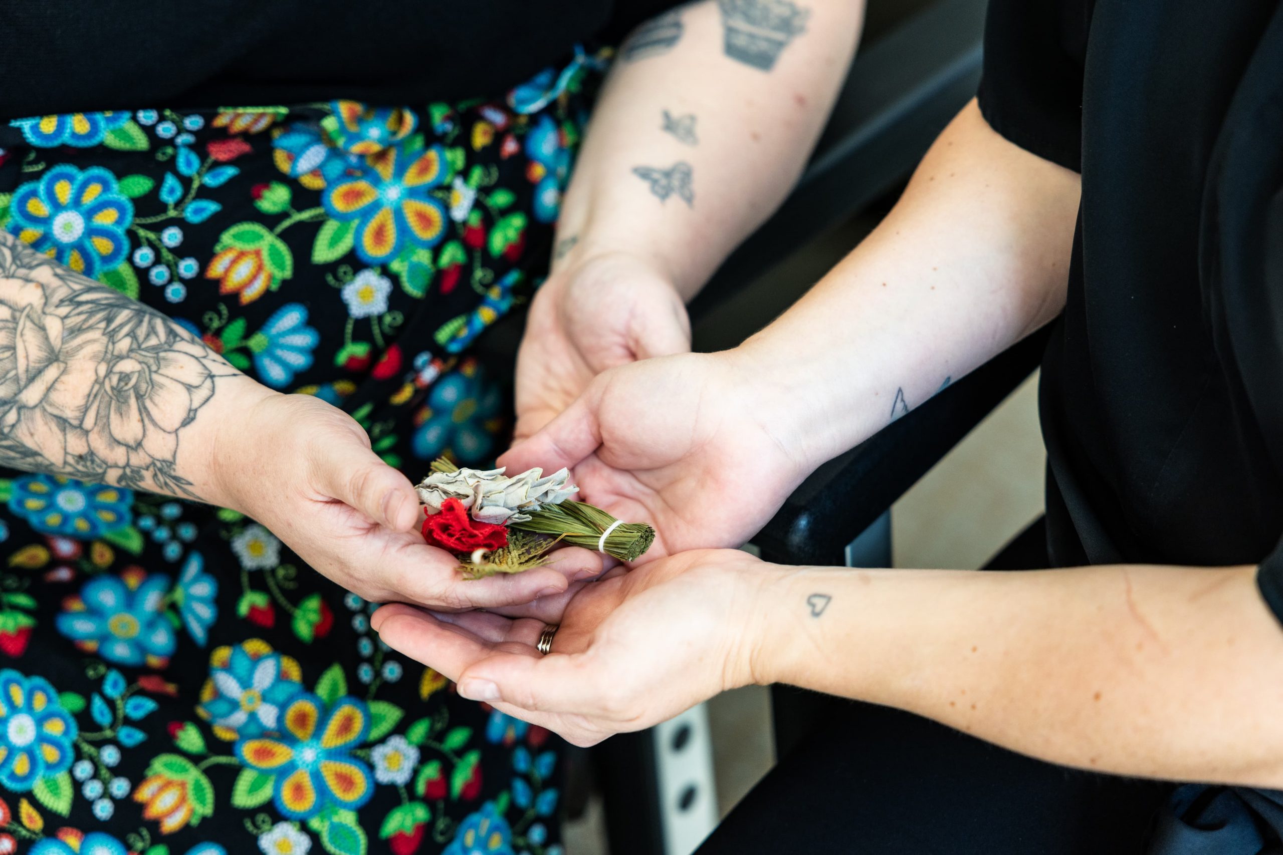 Two people with tattoos hold a small bundle of sage and a red flower together in their hands.