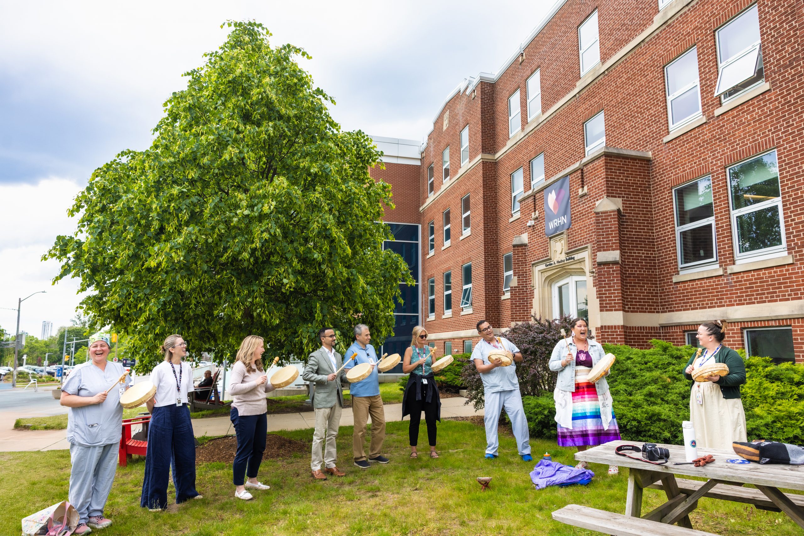 A group of people stand in a circle outside a brick building, holding hand drums and participating in an outdoor activity.