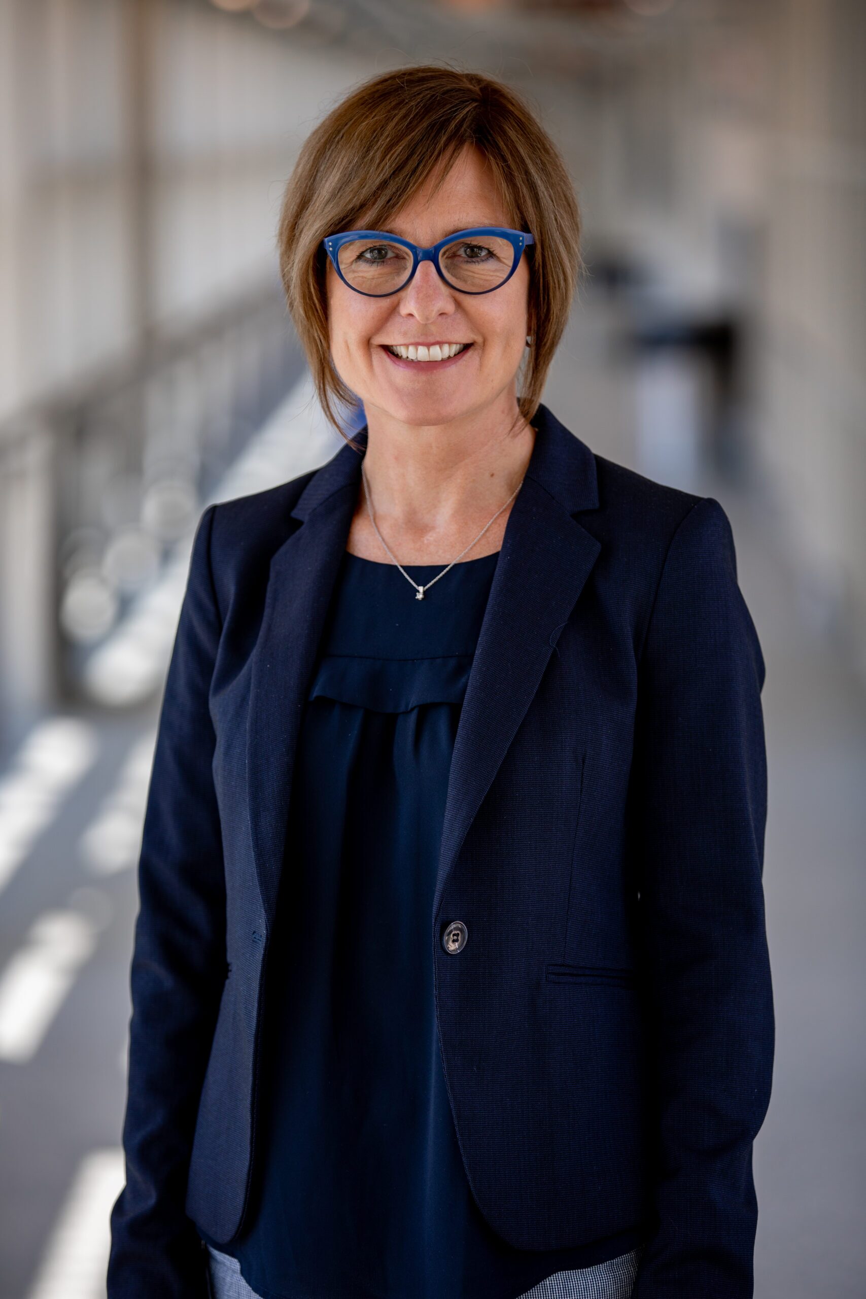 A woman with short brown hair and glasses smiles, wearing a black top and standing indoors.