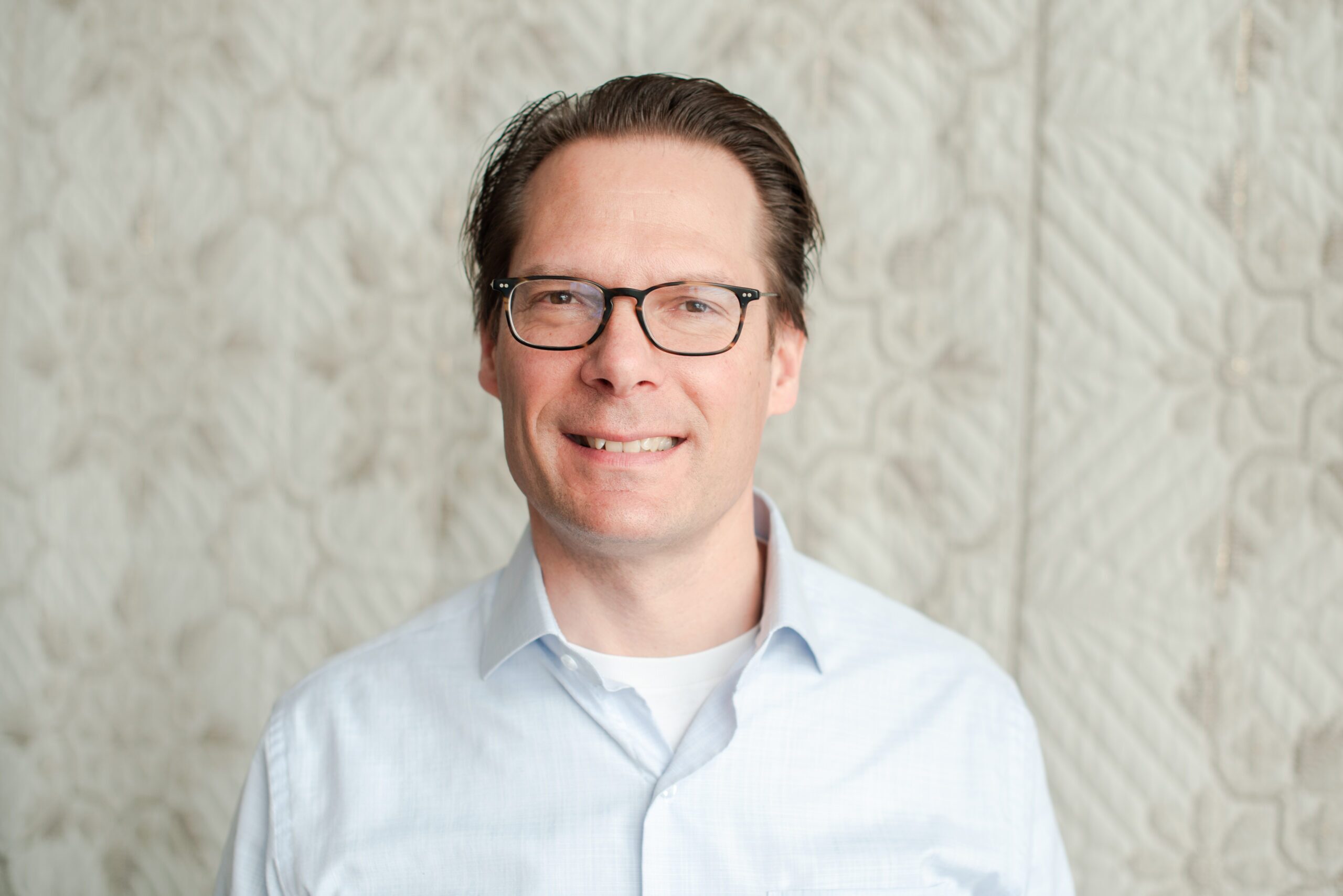 James wearing glasses and light brown hair wearing a light blue shirt, smiling in front of a patterned light background.