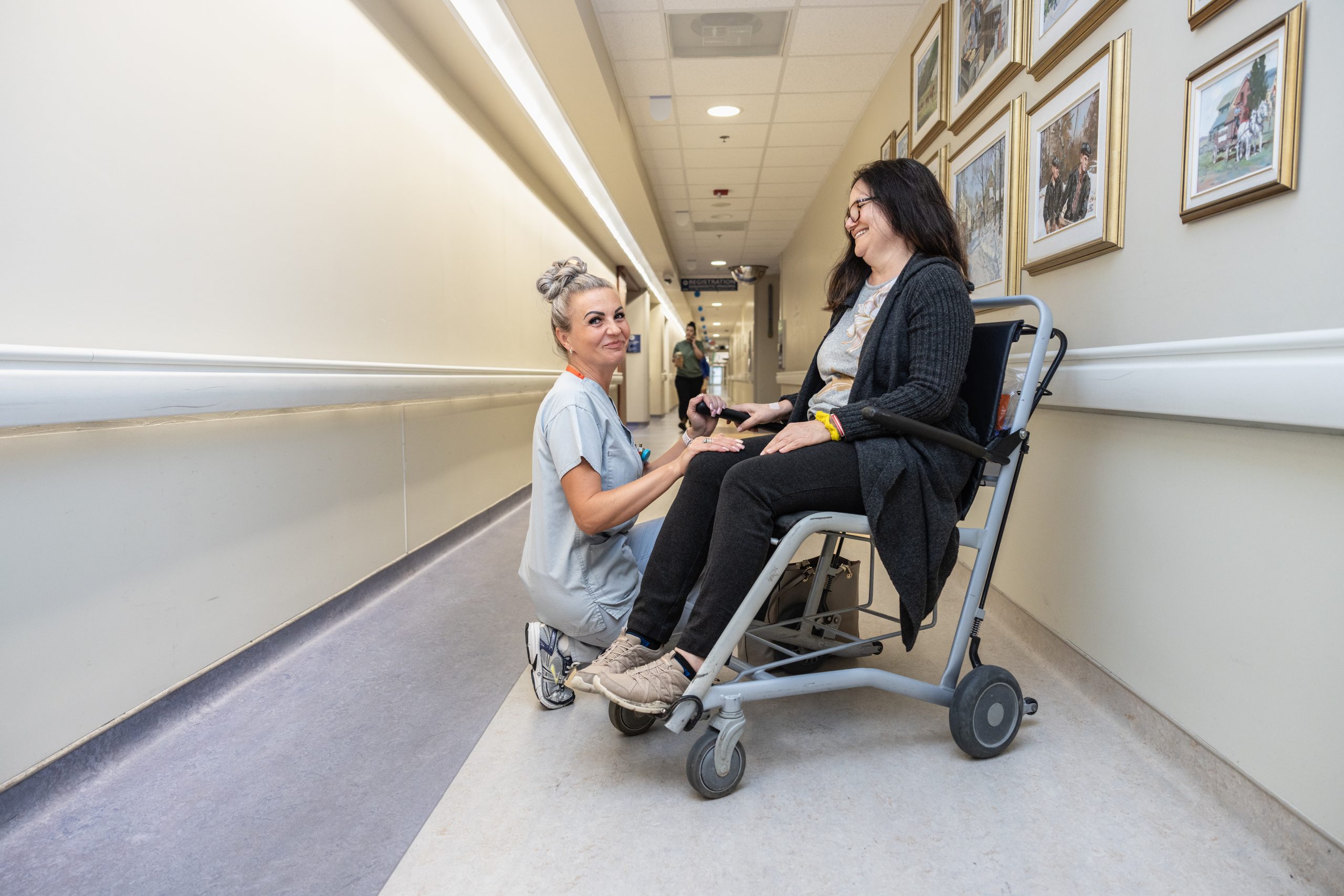 A nurse kneels beside a patient in a wheelchair, smiling and holding her hands in a hospital hallway.
