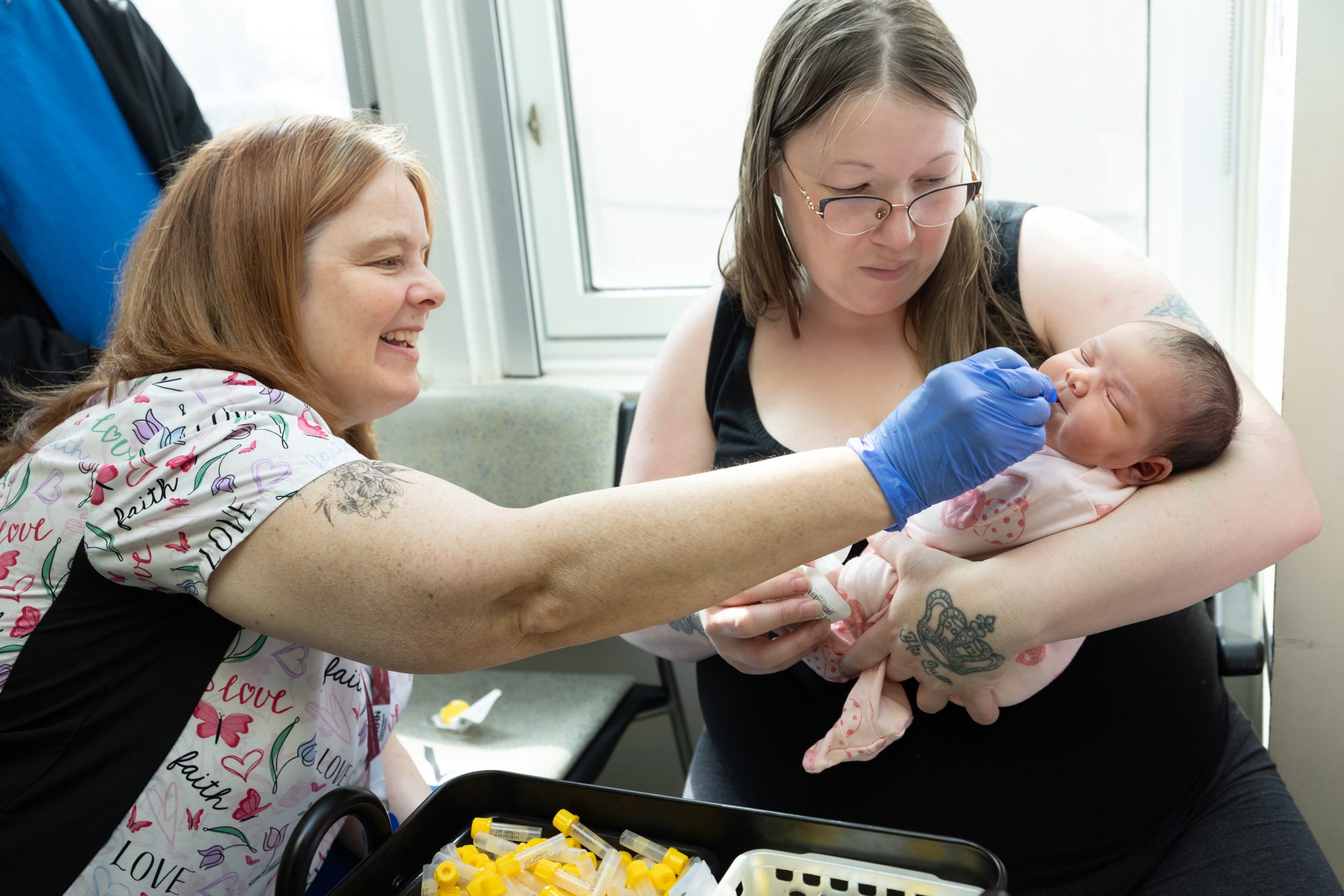 A nurse in scrubs takes a swab from a newborn's mouth as a woman holds the baby in a medical setting.