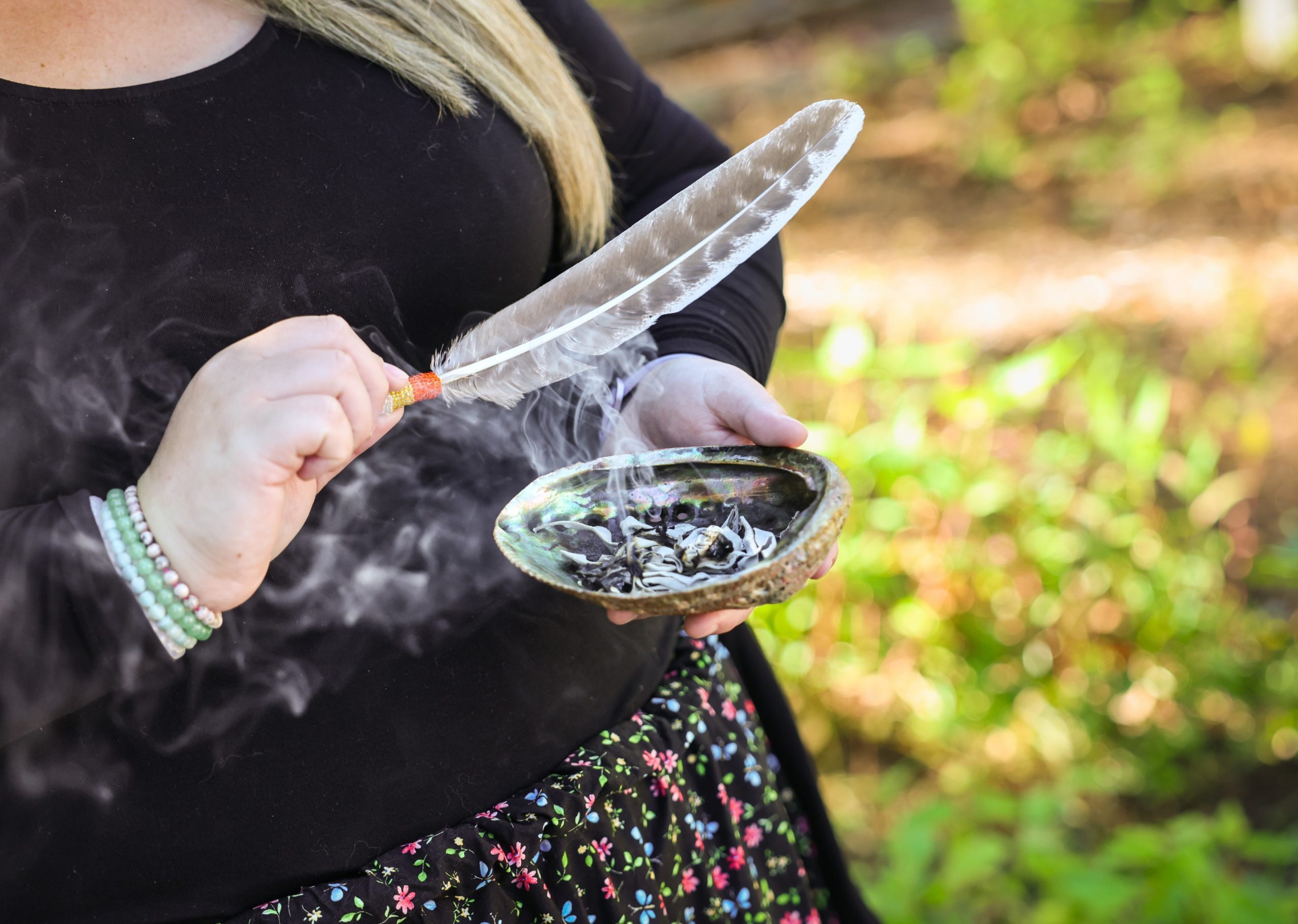Person holding a feather and a shell with smoldering herbs, creating smoke, outdoors with greenery in the background.