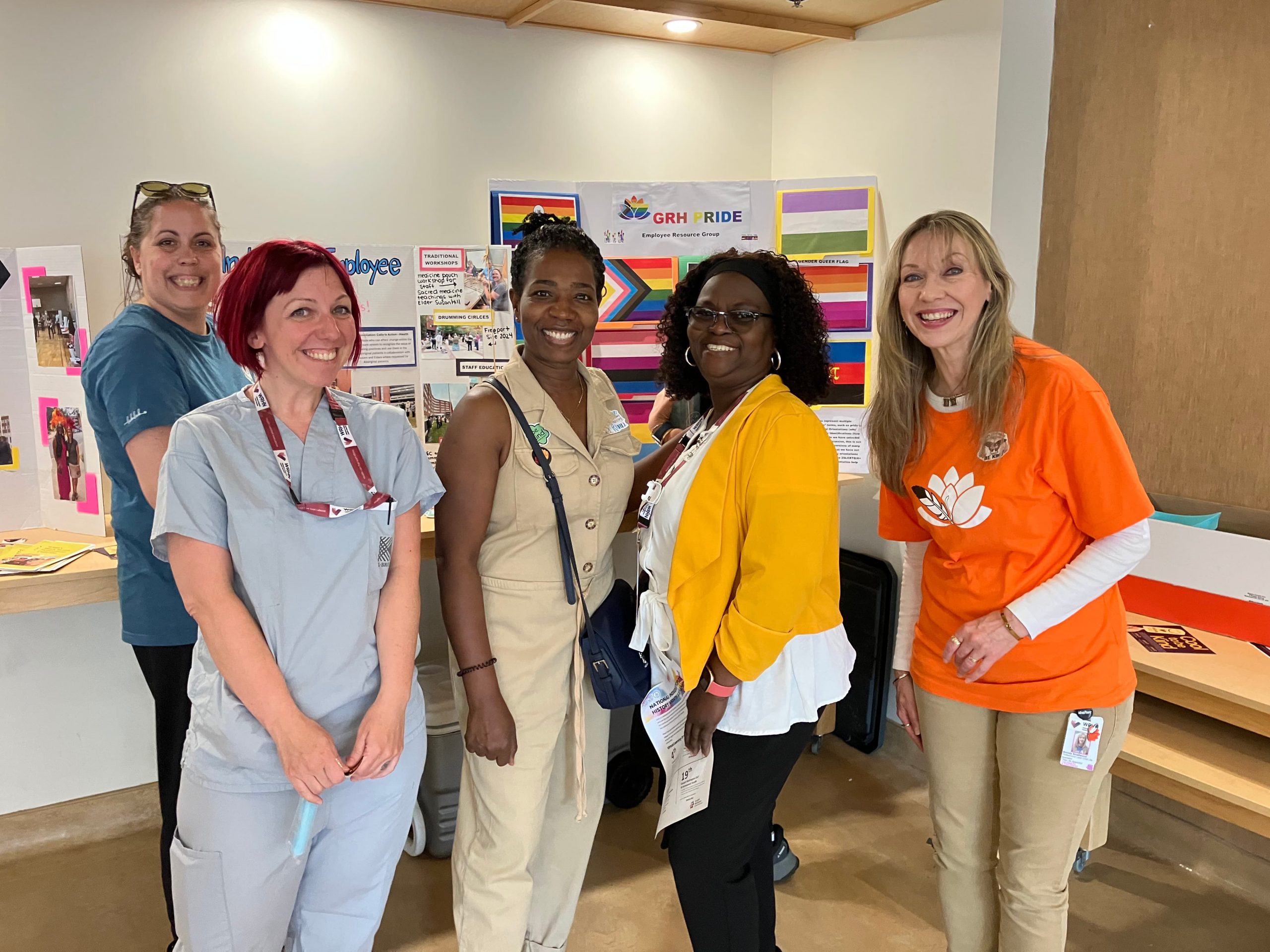 五名女性站在明亮房间里，面带微笑地伫立于展示板前，板上陈列着各式海报与彩虹旗。