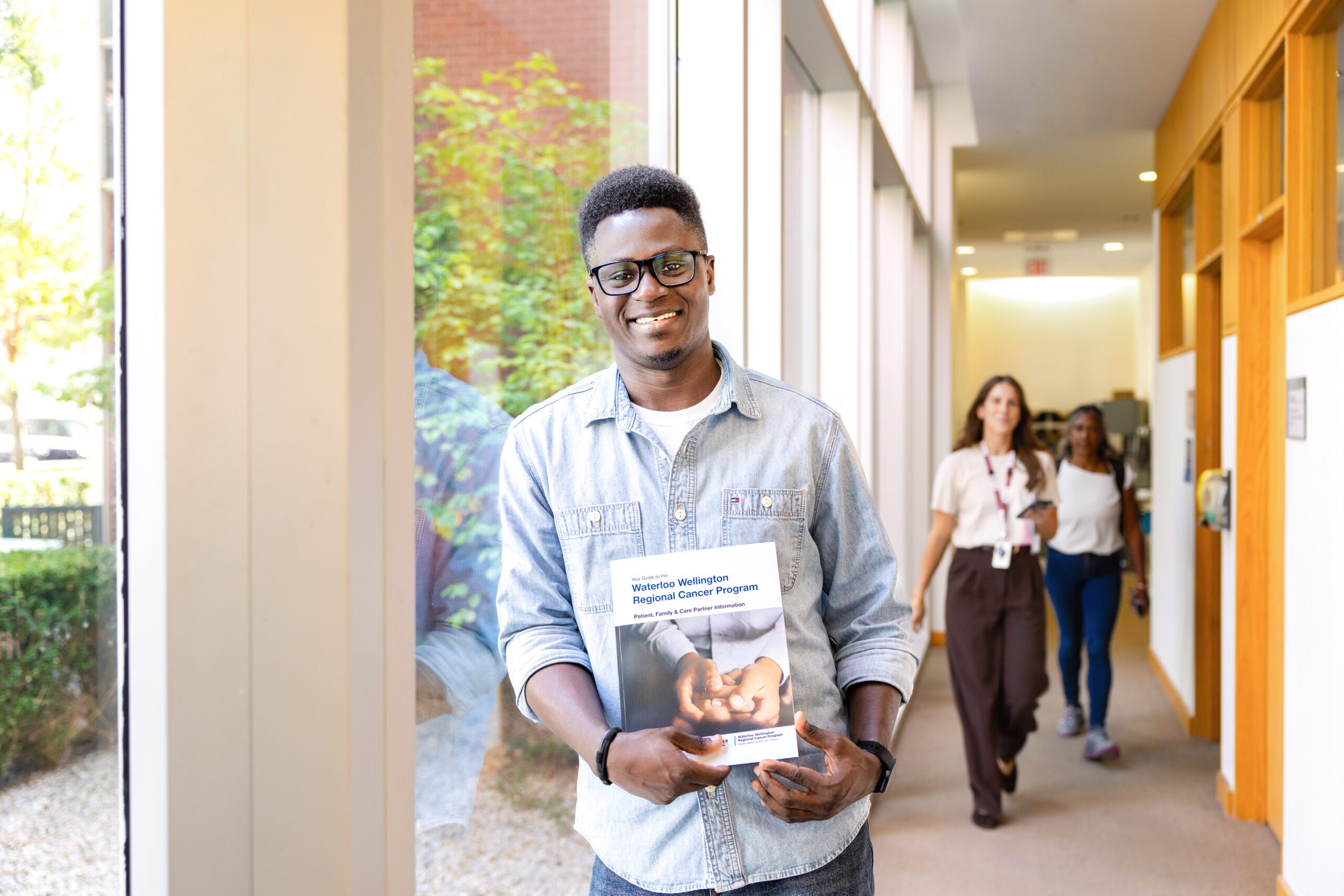 A man in glasses holds a brochure and stands in a hallway with two women walking in the background.