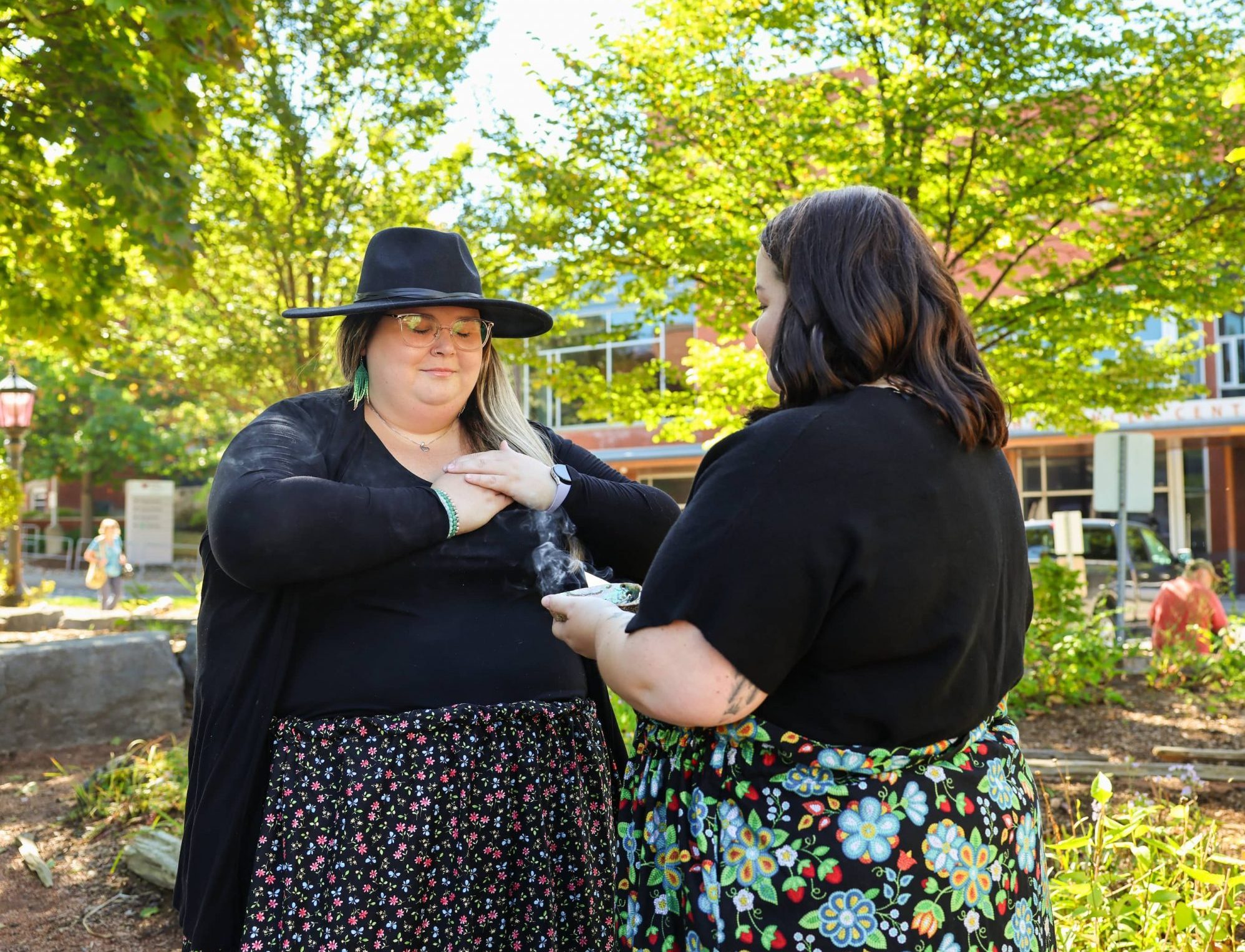 Two people standing outside smudging with sacred medicine. Trees and a hospital building are in the background.