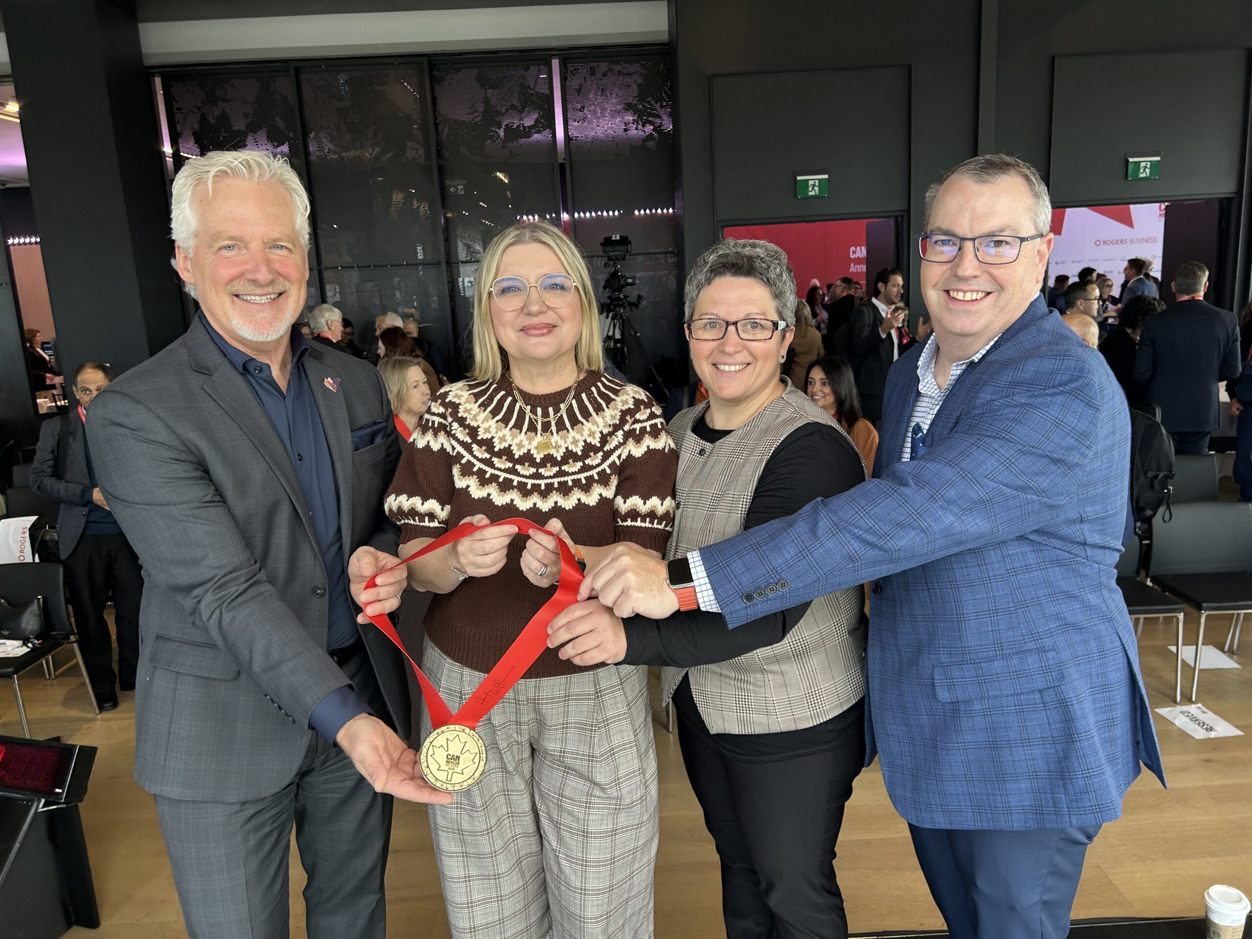 Four people standing indoors, holding a large gold medal together, smiling at the camera during an event.