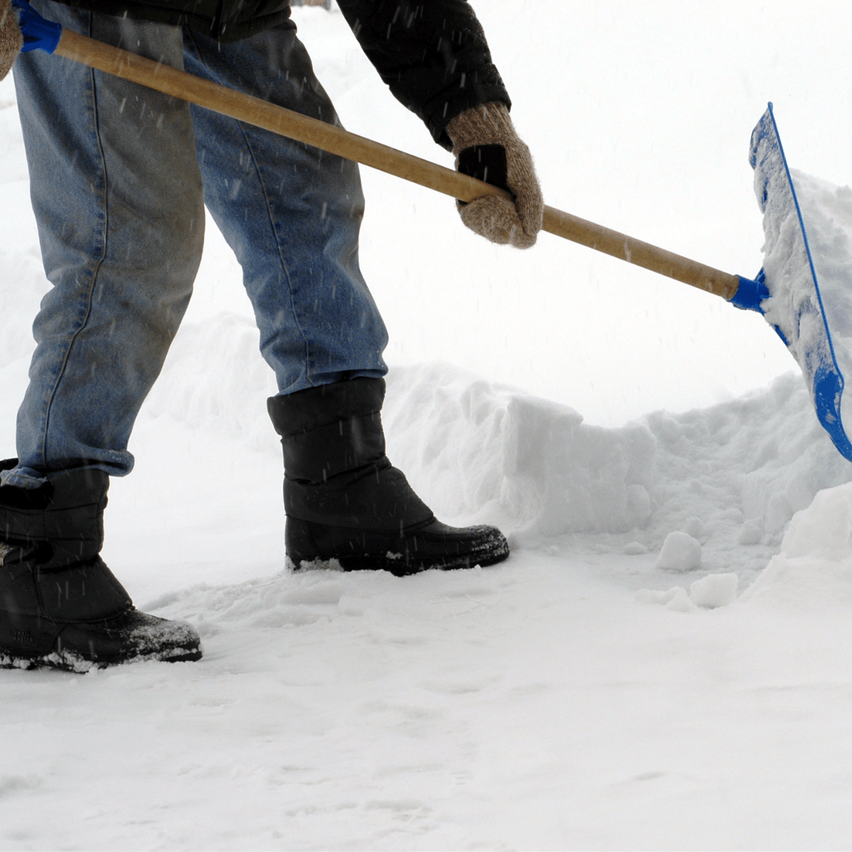 Person wearing winter boots and gloves uses a blue snow shovel to clear snow from the ground.