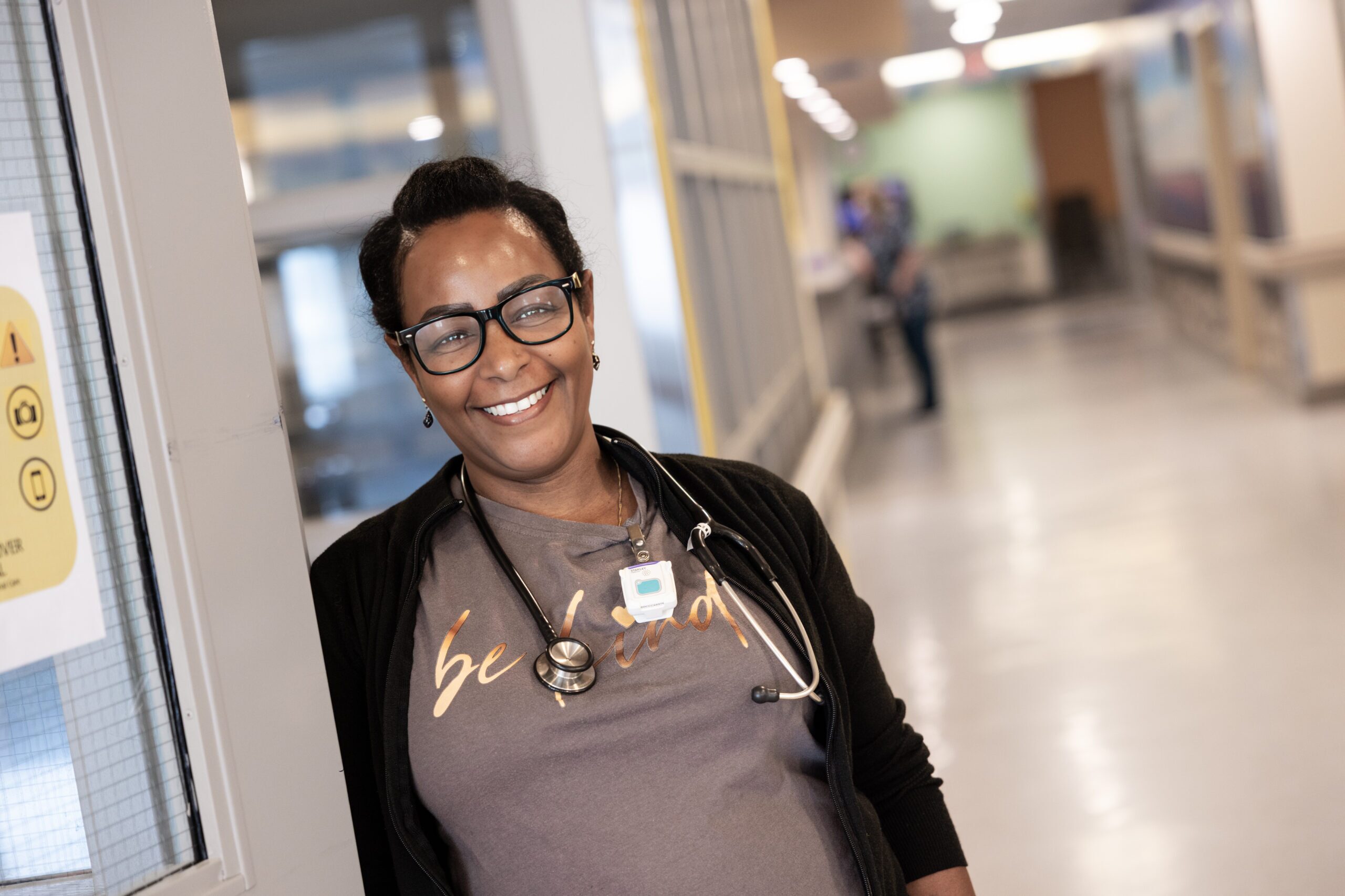 A nurse leans against the wall in one of the units, smiling. They have a stethoscope around their neck and wear a shirt that says, “Be Kind.”