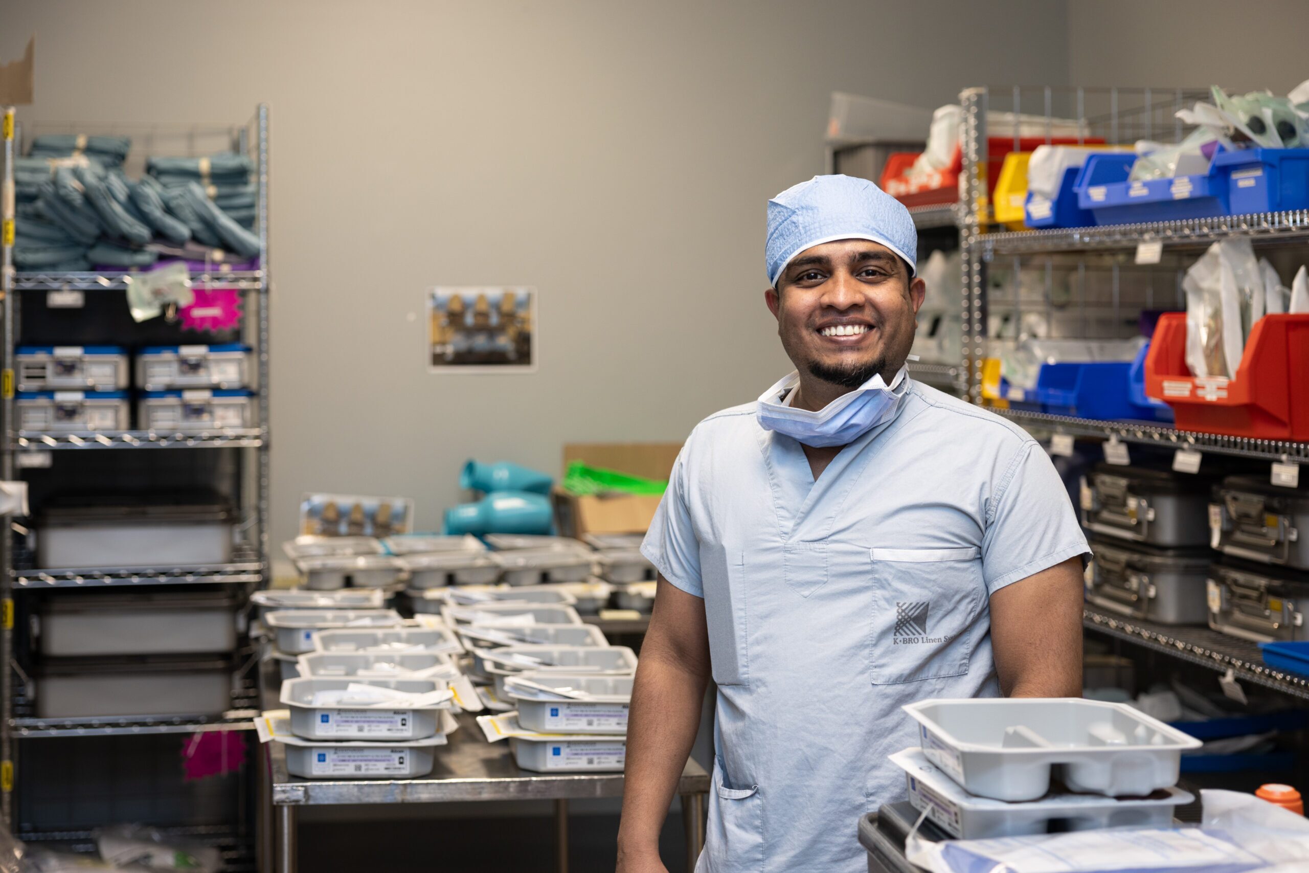 A team member in blue scrubs and a scrub cap stands in a supply room, smiling.