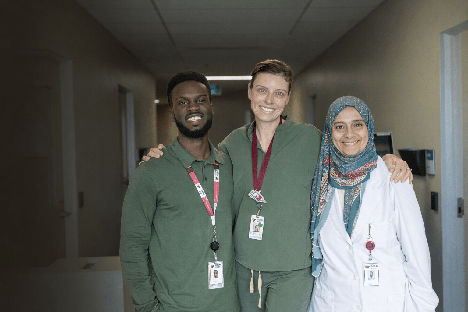 Three team members stand together in a hallway with their arms around each other, smiling.