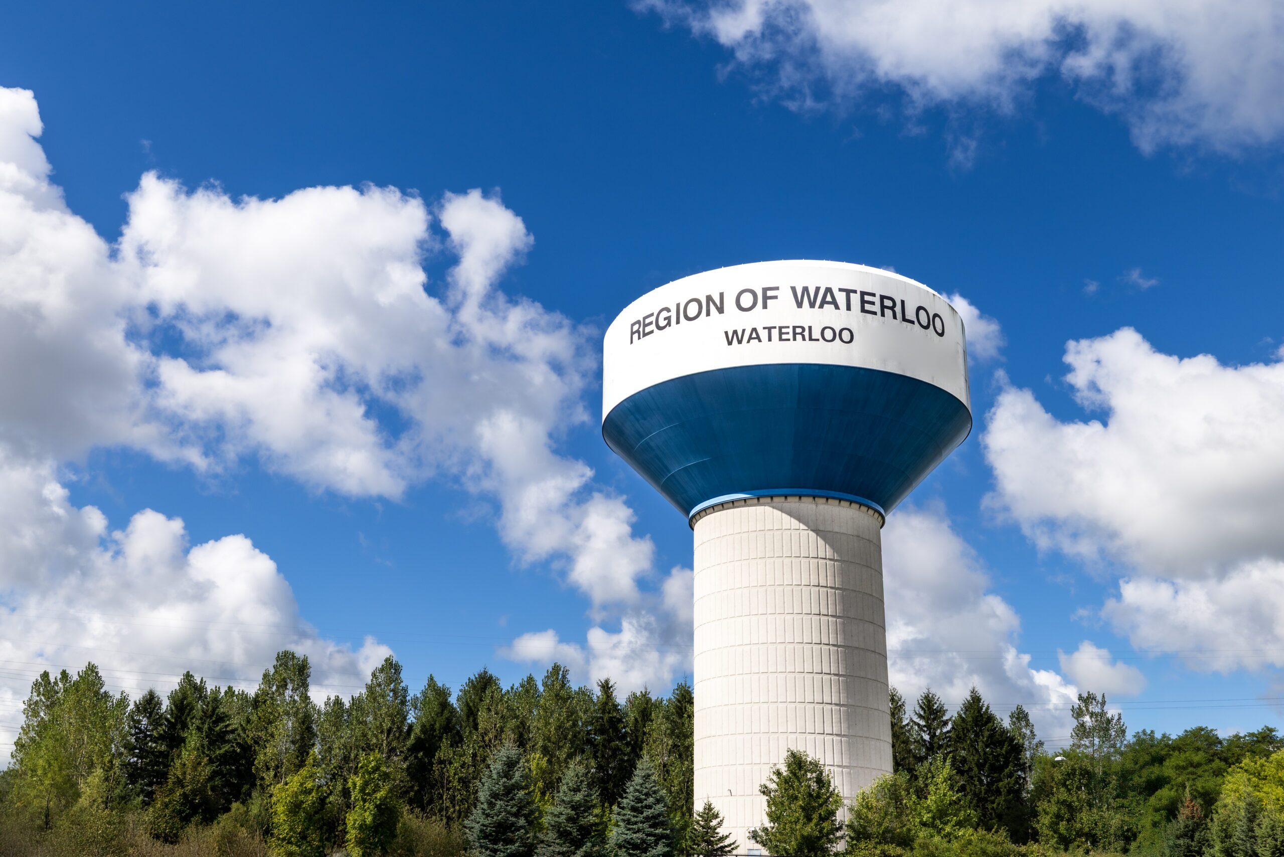A Region of Waterloo water tower stands above the tree line against a blue sky with white clouds.