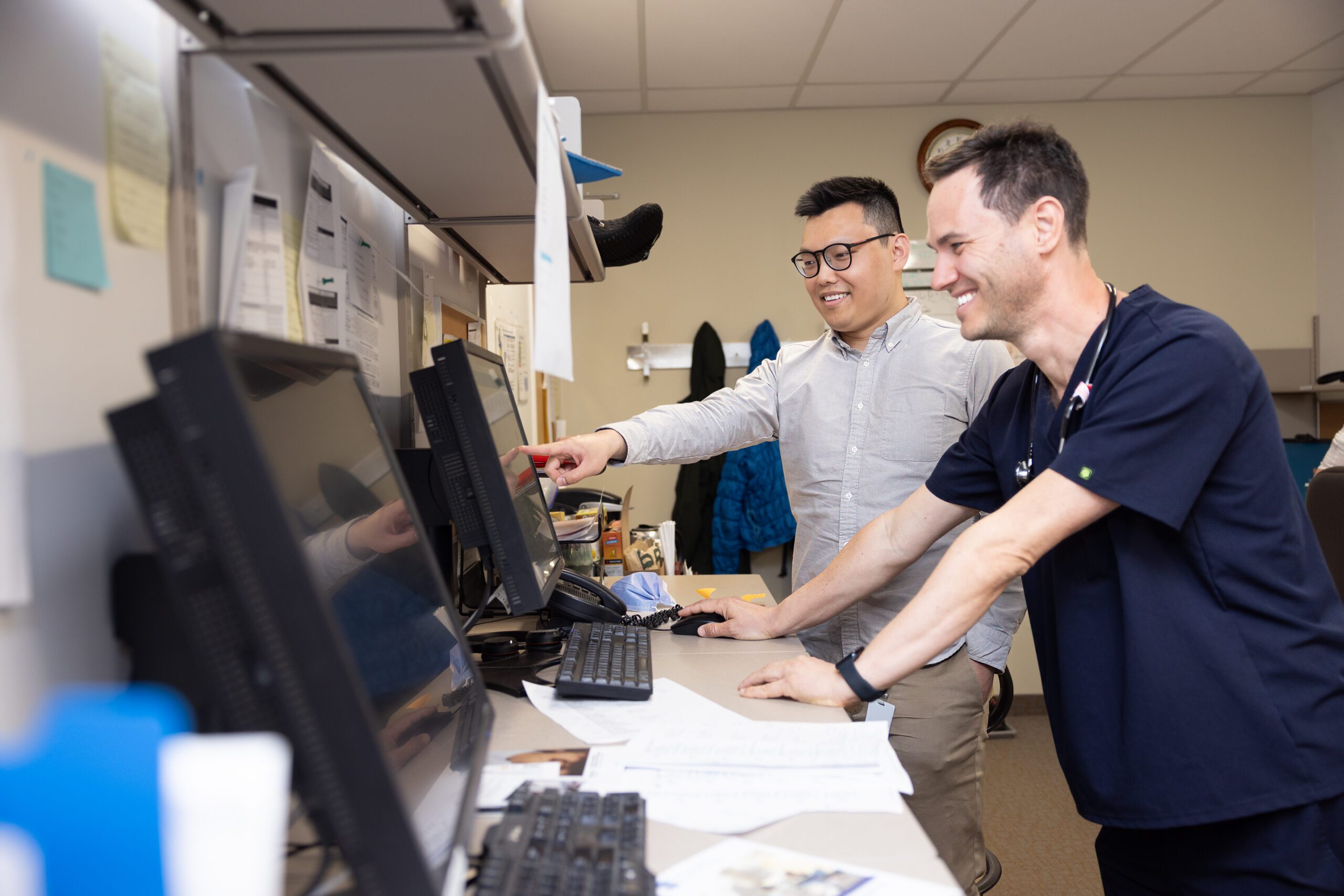 Two team members look at information on an office computer together. One is pointing at the screen and the other is smiling.