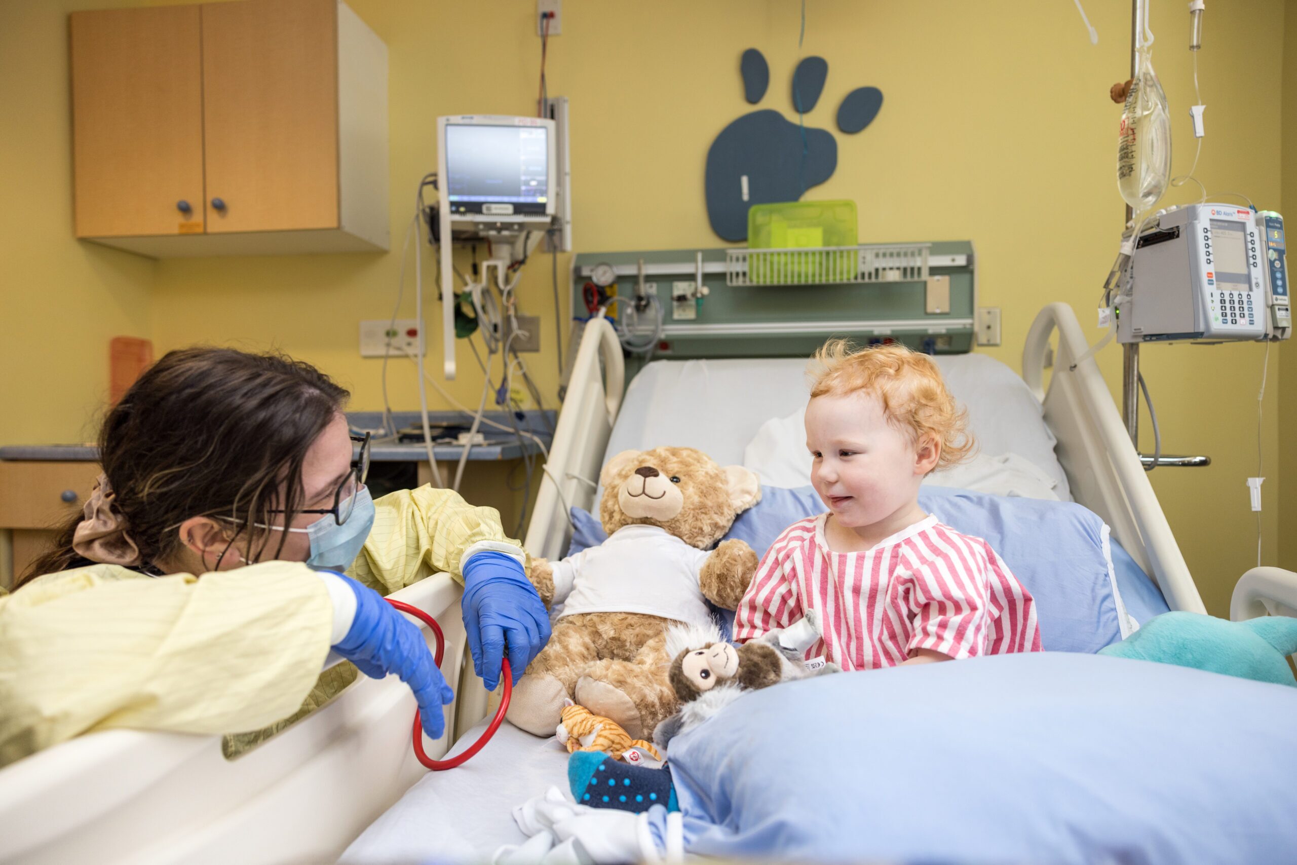 A toddler sits with stuffed toys in a hospital bed. A team member is down at the child’s eye level talking to them.