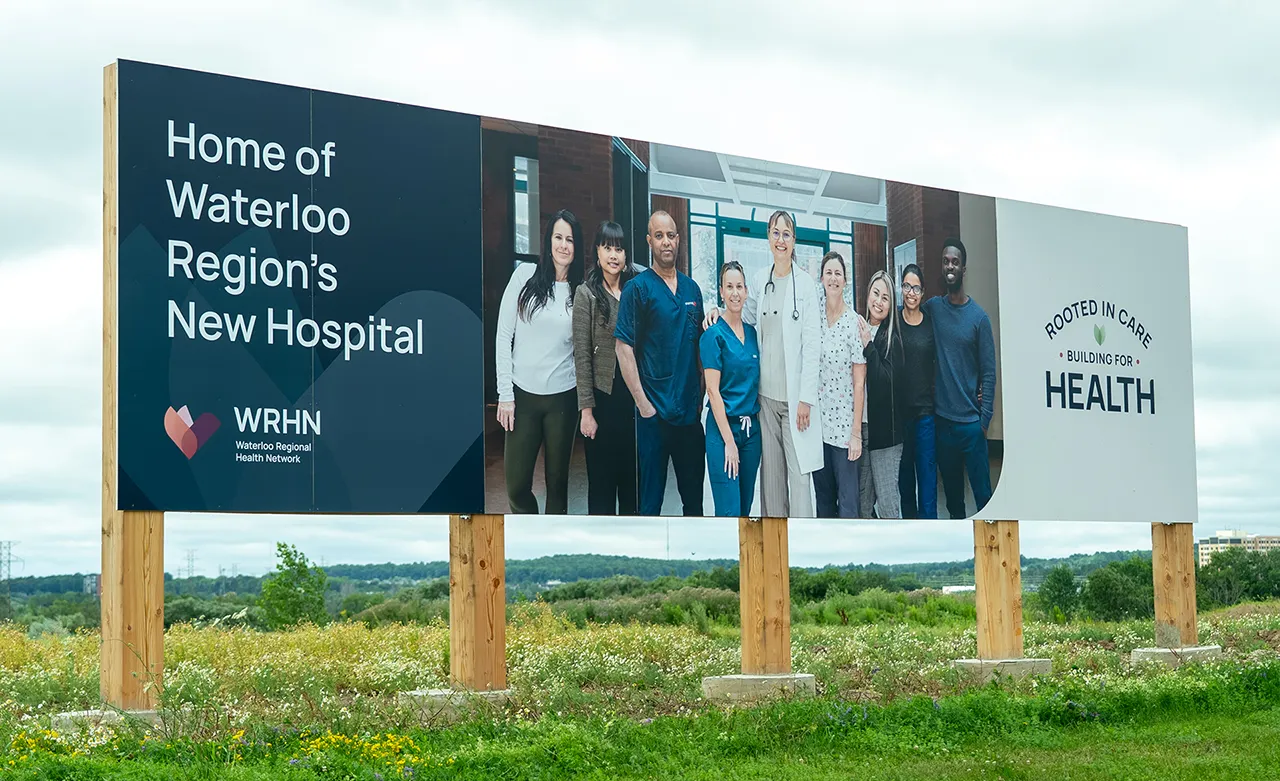 A sign on the site of Waterloo Region’s new hospital. It includes a photo of diverse team members with the text, “Home of Waterloo Region’s New Hospital,” and the tagline, “Rooted in Care. Building for Health.”