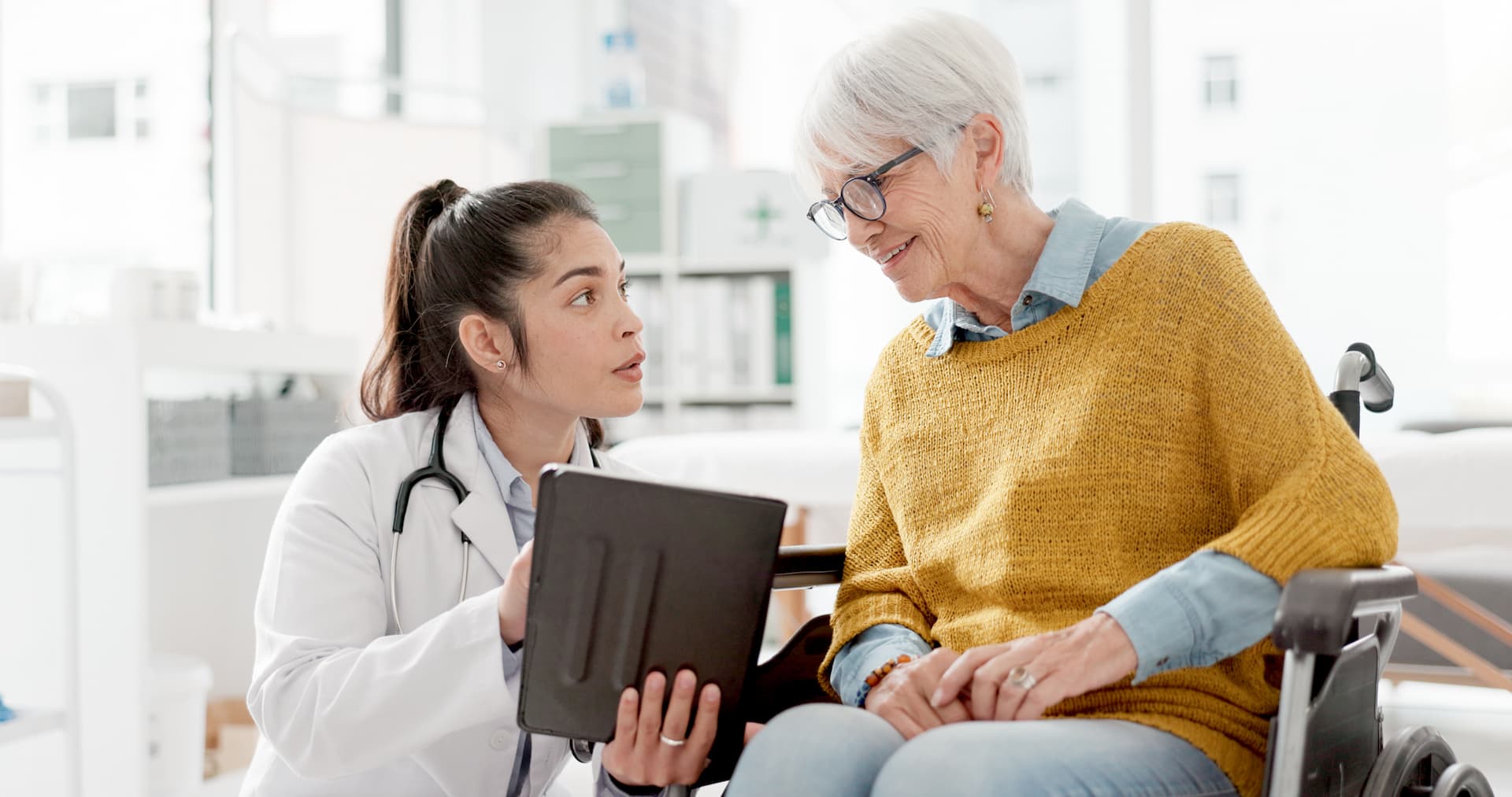 Young doctor shows information on a tablet to an older woman in a wheelchair during a medical consultation.