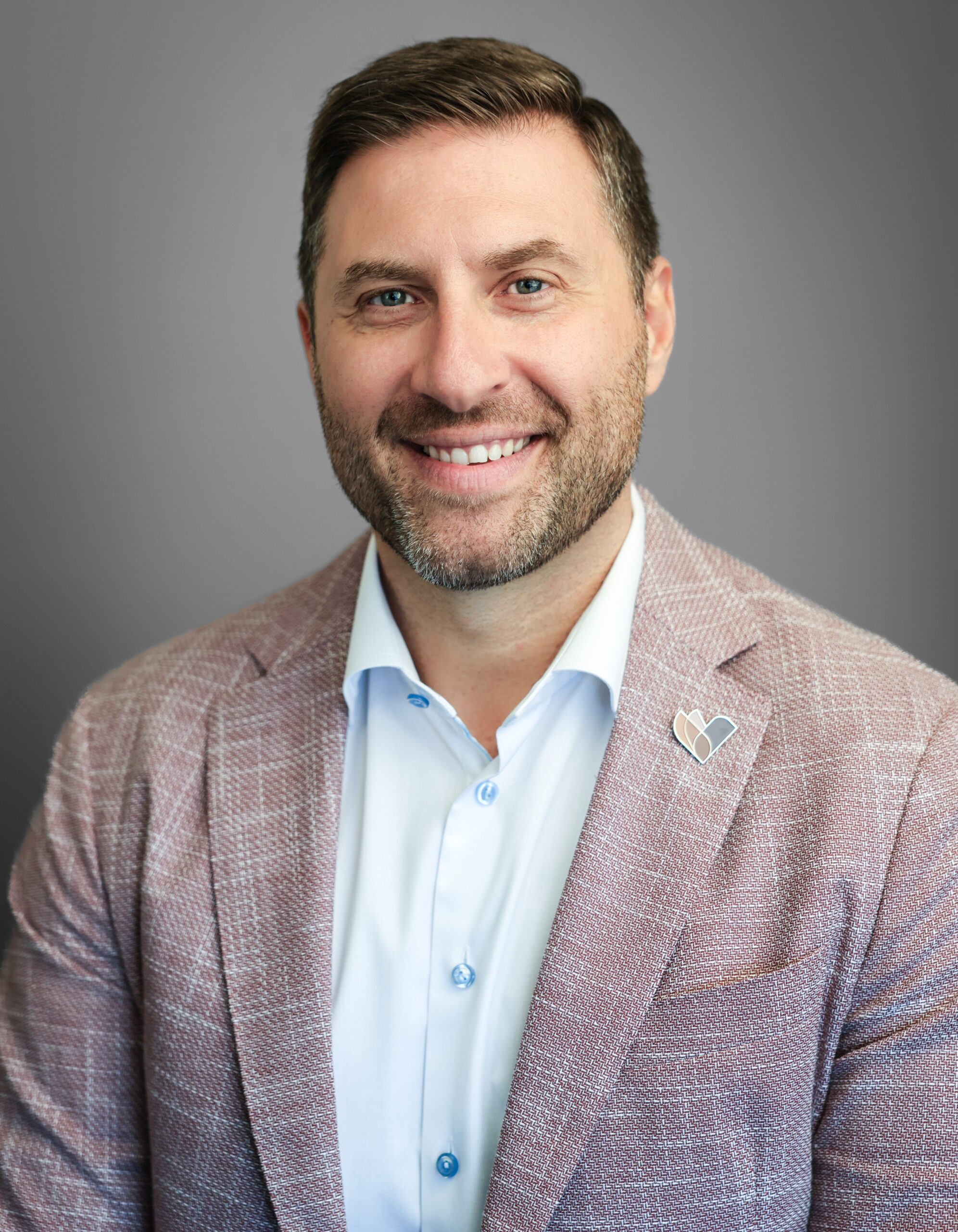 Man wearing a light blue shirt and pink blazer, smiling in front of a plain gray background.