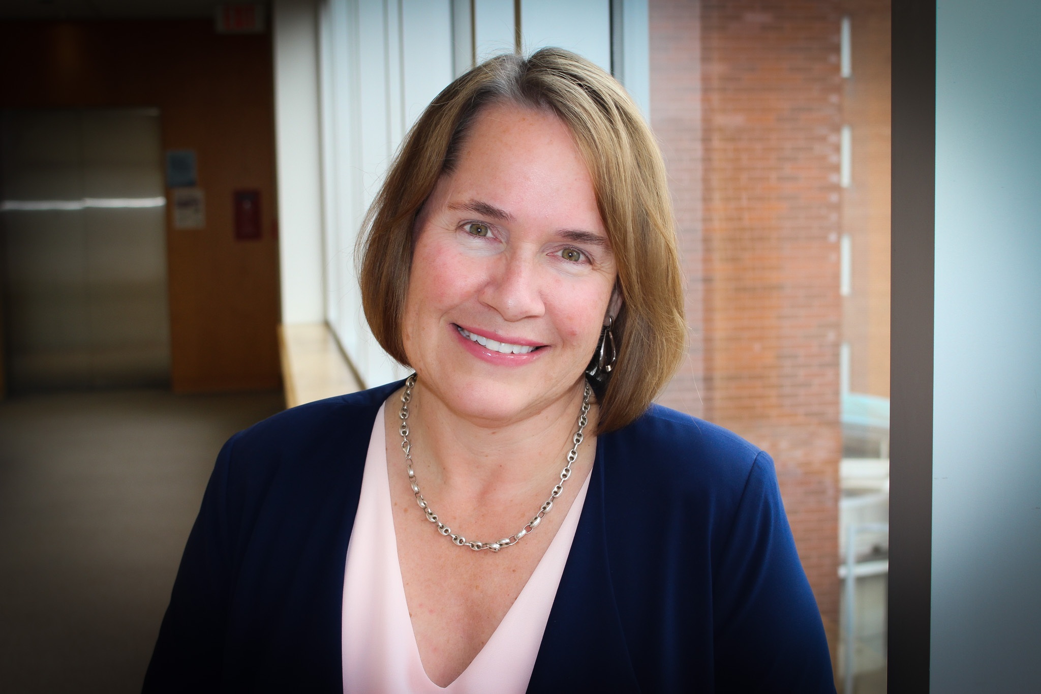 Liz smiles with short brown hair in a blue blazer and pink top stands by a window, smiling at the camera.