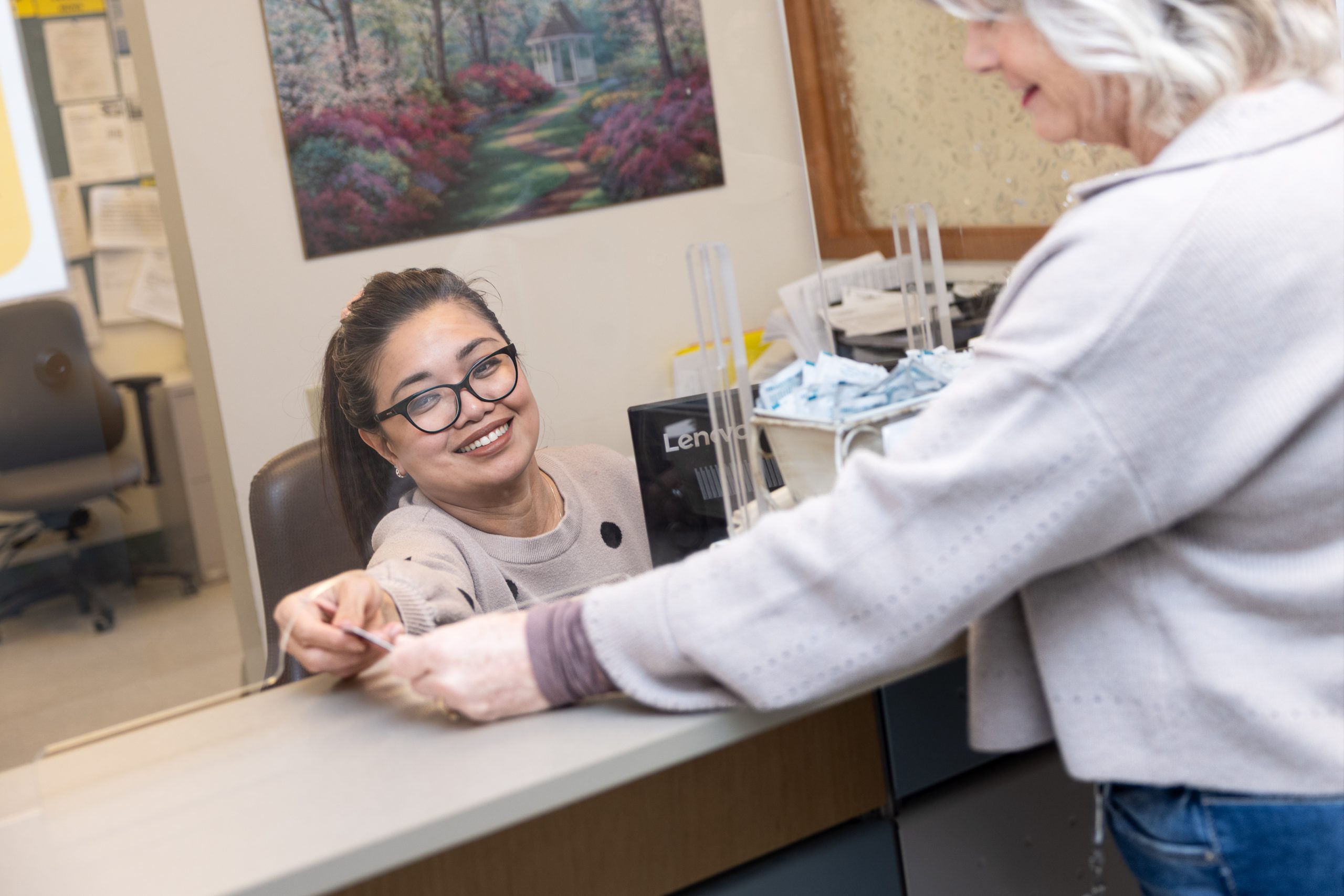 A receptionist behind a counter smiles and accepts a card from a woman standing on the other side.