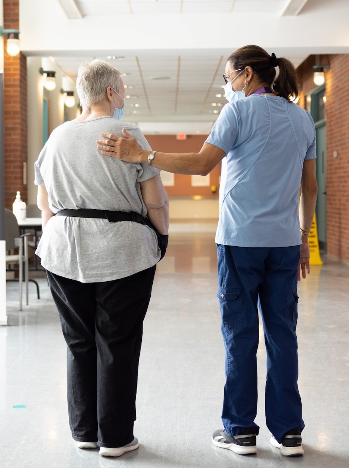 A healthcare worker stands beside an older adult, gently touching their shoulder as they walk down a hallway together.