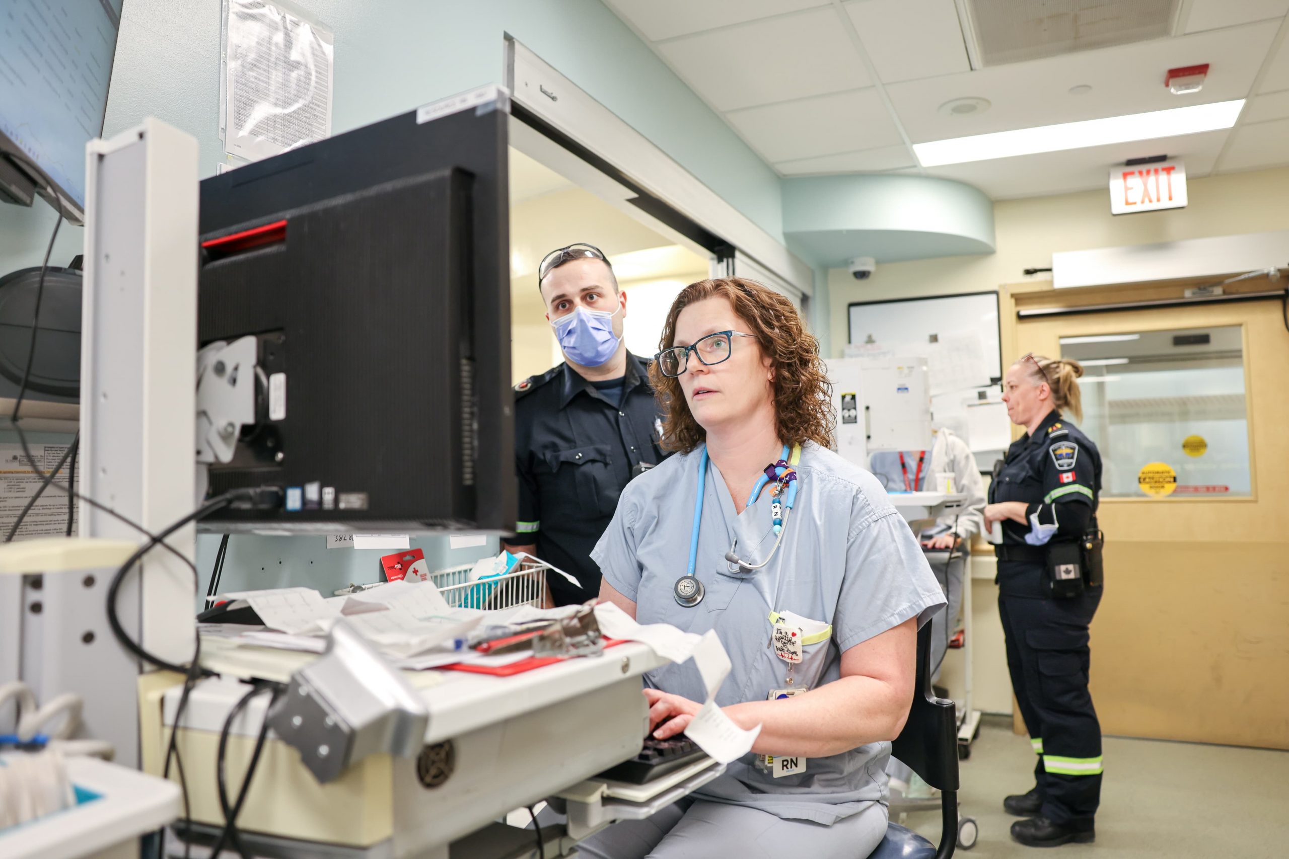 A nurse works at a computer station in a hospital as two paramedics stand nearby in uniform.