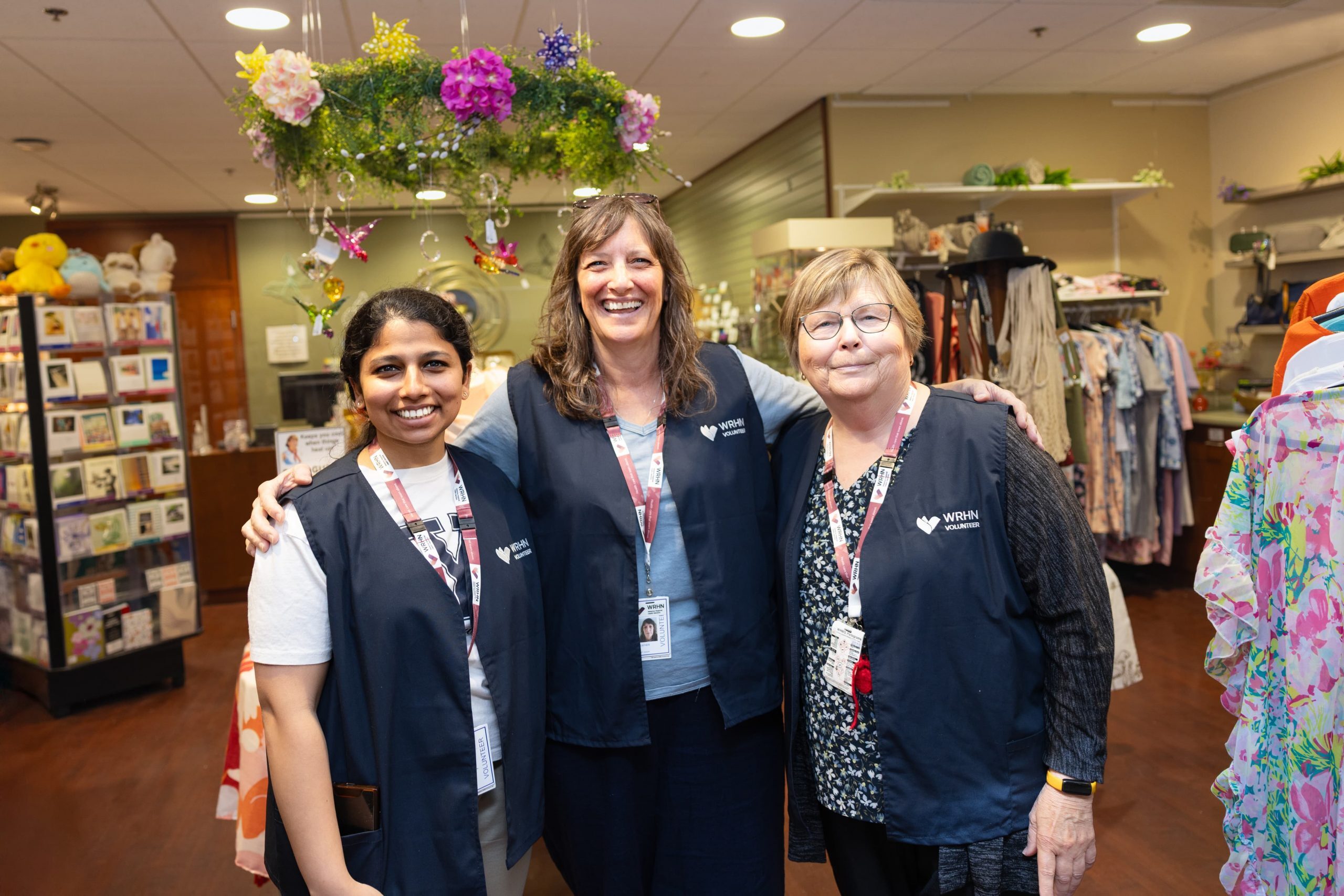 Three women wearing navy vests stand together and smile inside a gift shop with clothing and greeting cards.