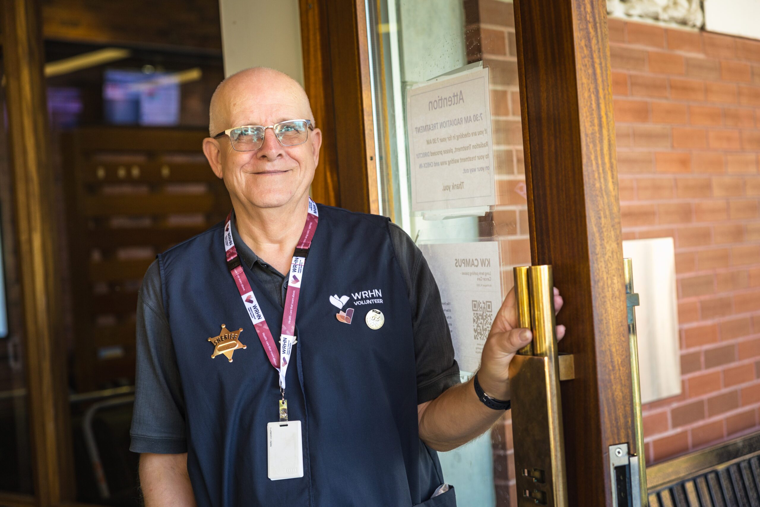 Two women wearing volunteer vests and name badges stand in front of a staircase inside a building.