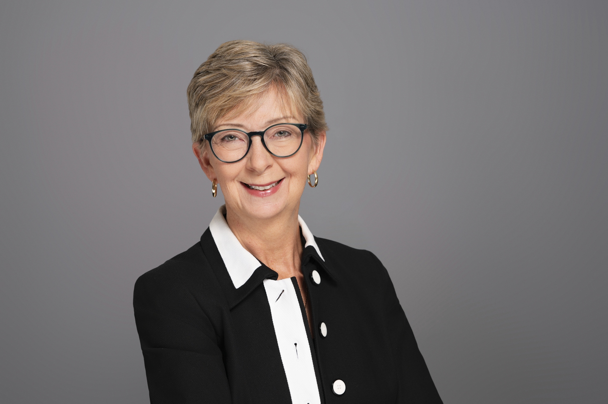 Janet smiles with short hair, glasses, and a black-and-white blazer poses against a plain grey background.