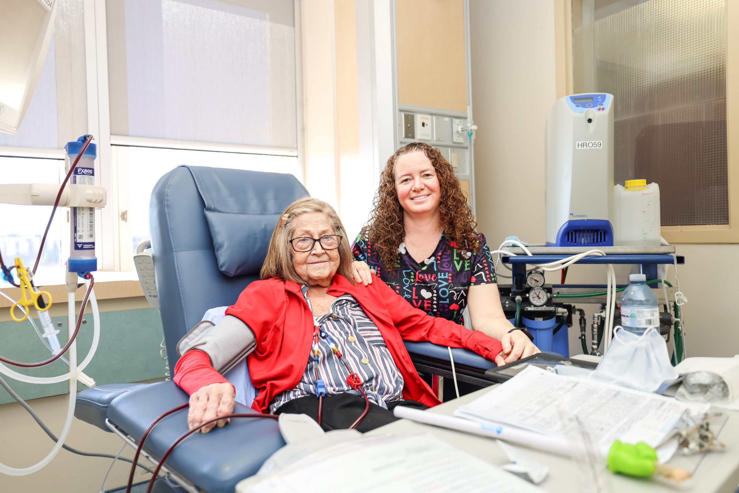 An elderly woman sits in a medical chair receiving IV treatment, with a nurse smiling beside her in a hospital room.