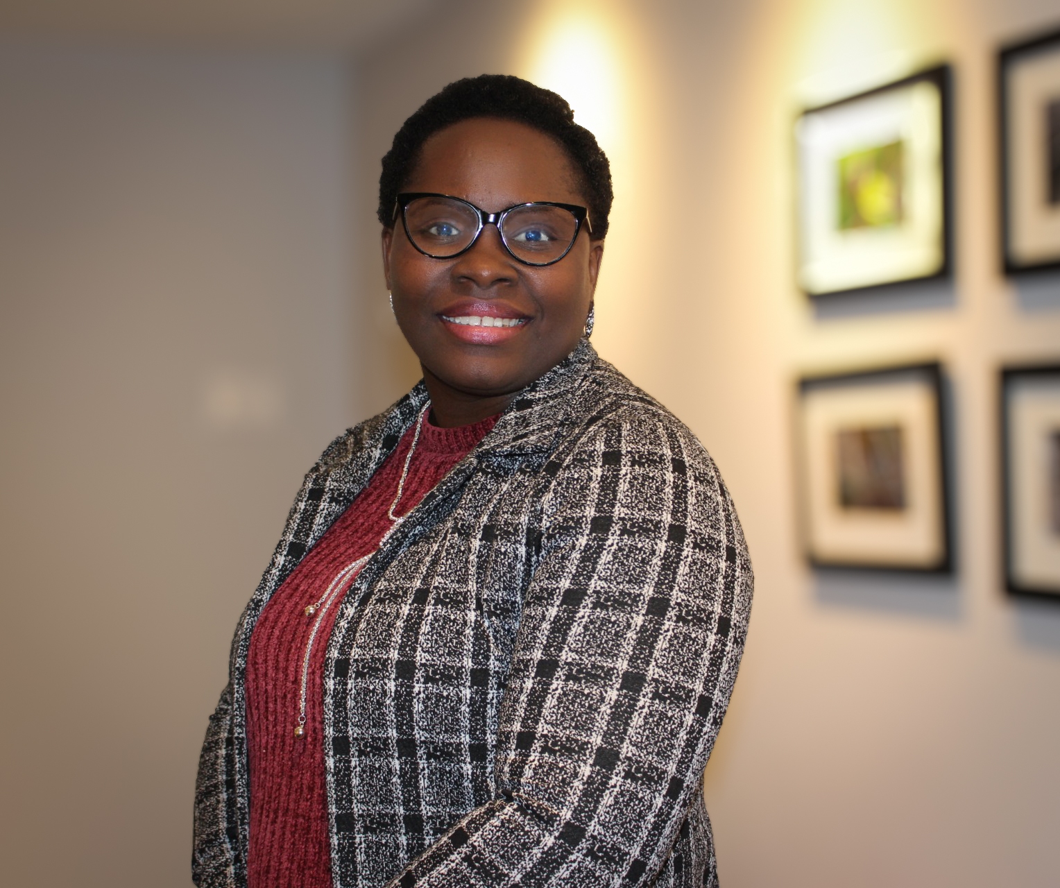 A woman wearing glasses, a plaid jacket, and a red top stands smiling in a hallway with framed pictures on the wall.