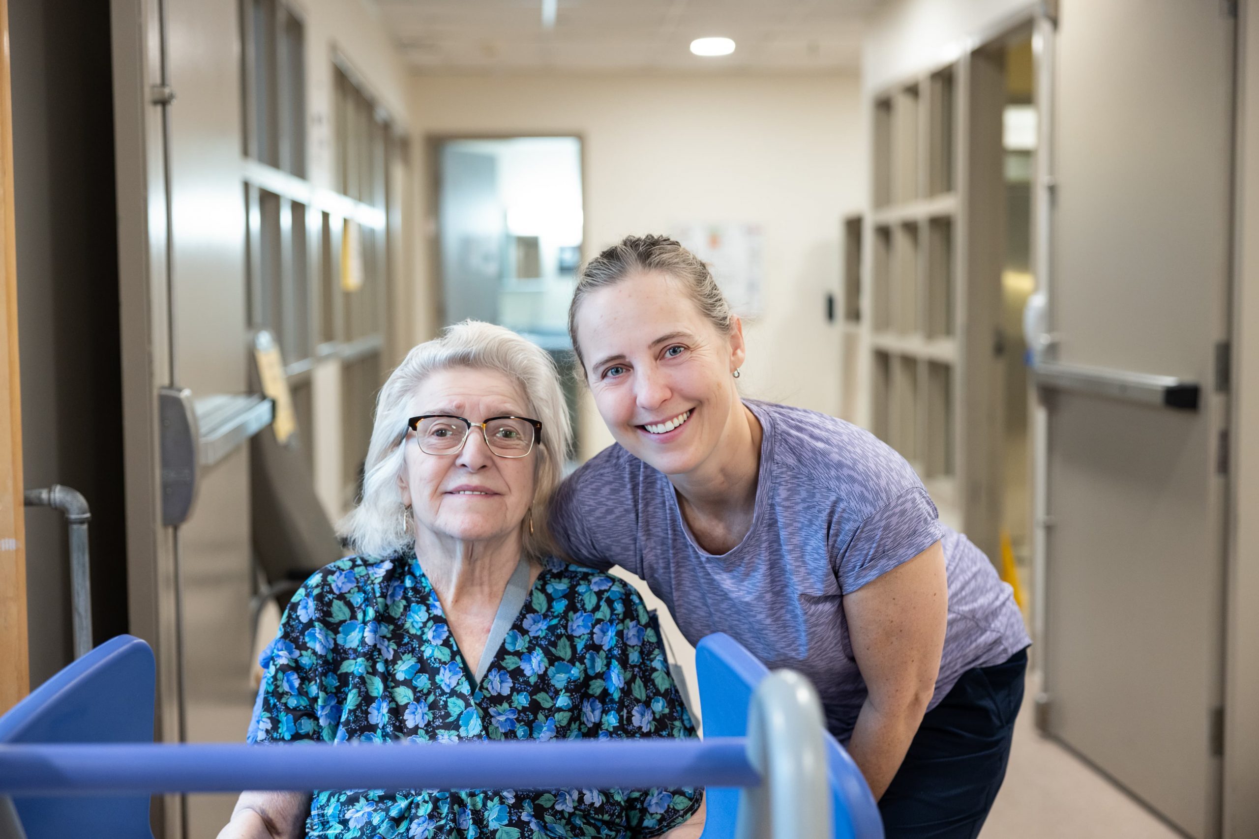 An older woman sits in a wheelchair next to a younger woman in a hallway, both looking at the camera.
