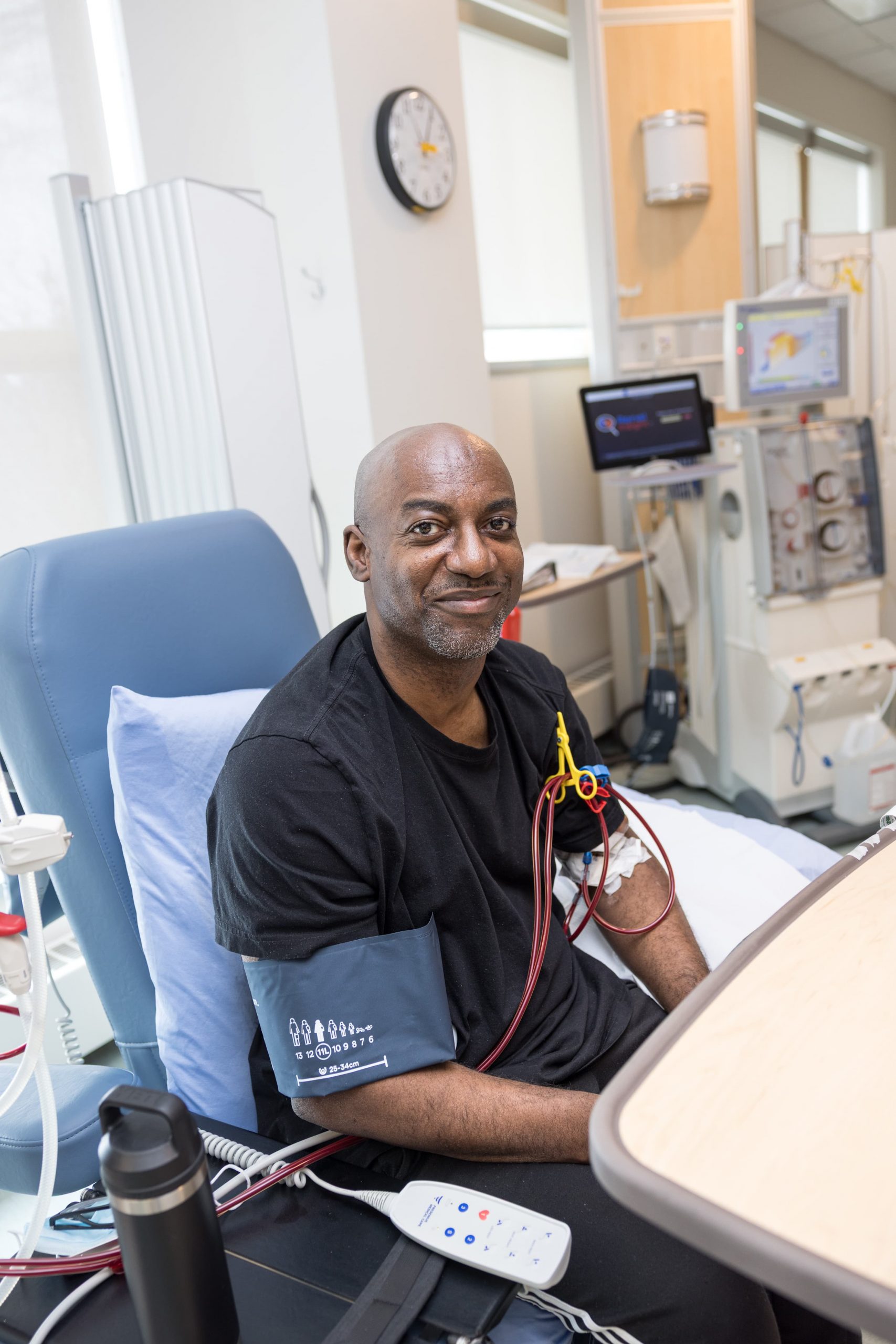 A man sitting in a medical chair receives dialysis treatment in a hospital room, connected to medical equipment.