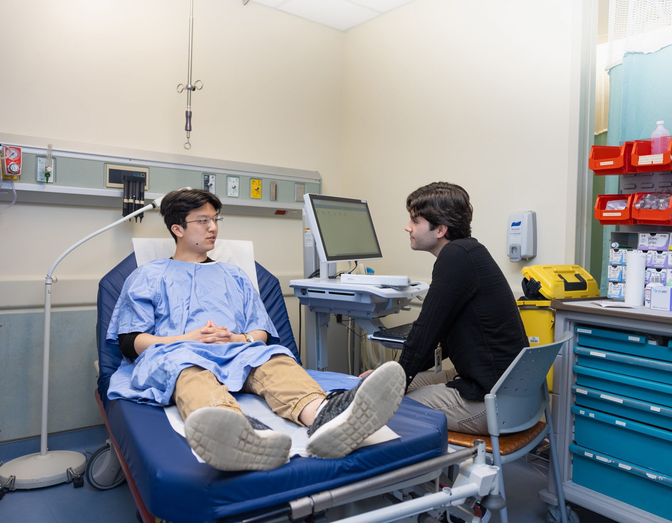 A patient in a hospital gown sits on a bed talking to a healthcare professional in an exam room.