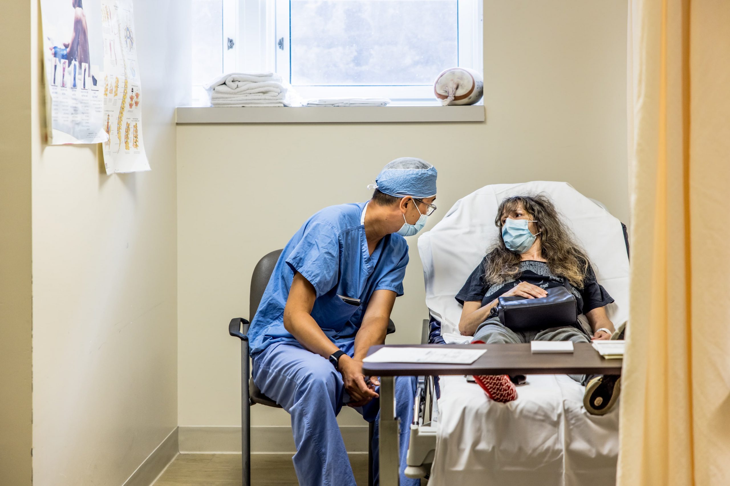 A healthcare worker in scrubs talks to a masked patient lying in a hospital bed in a medical room.