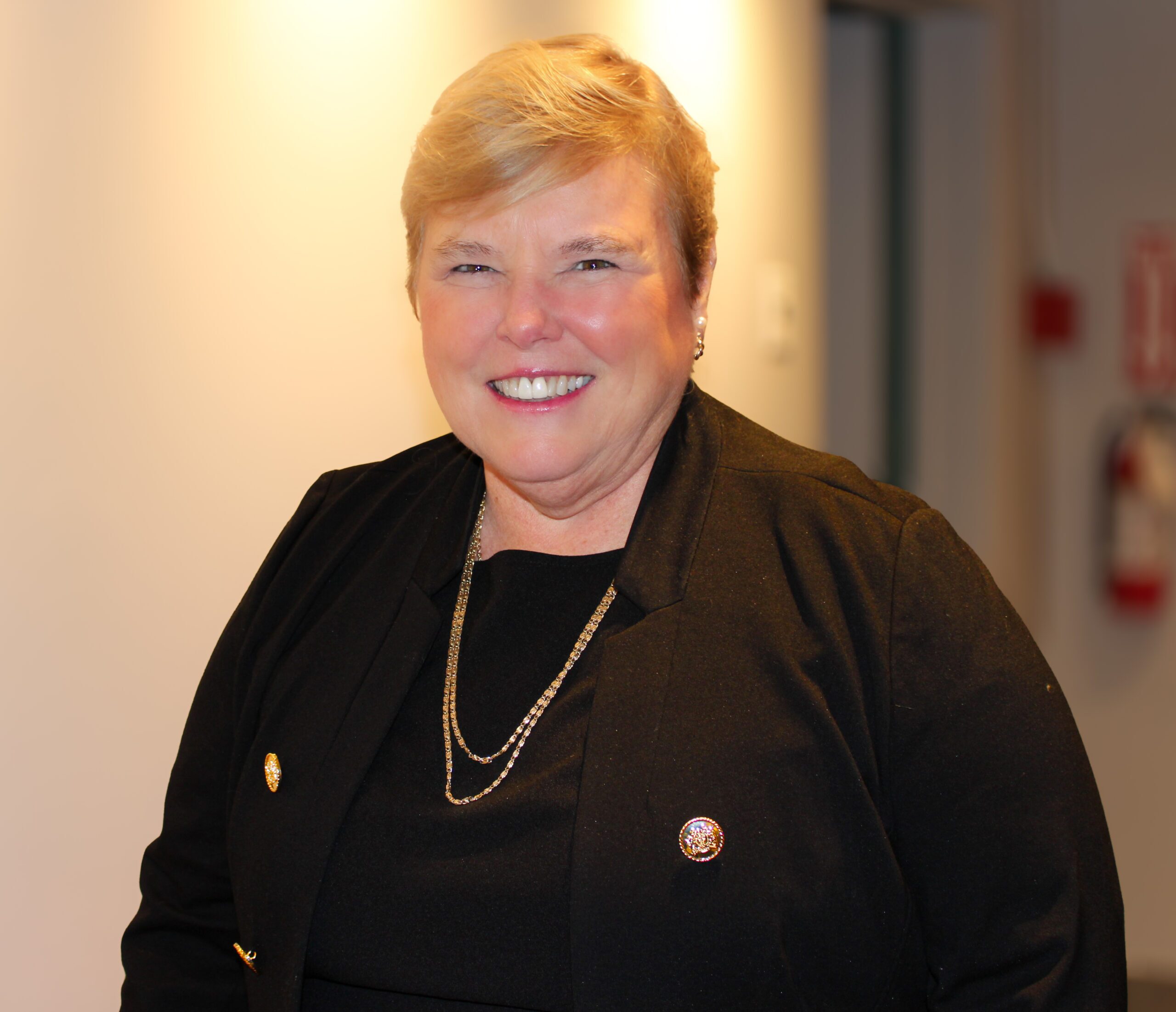 Sandra smiling with short blonde hair wearing a black blazer and gold necklace, standing indoors.