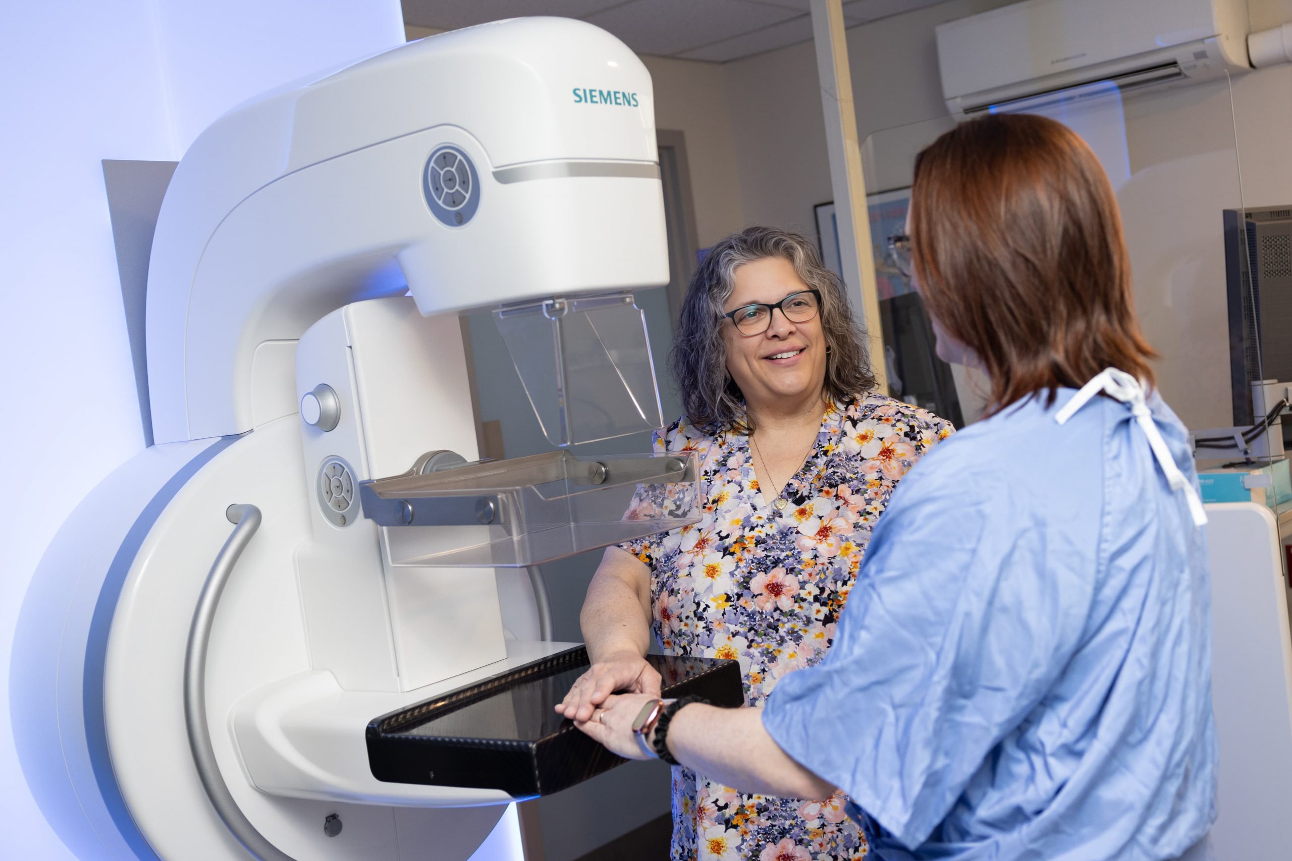 A healthcare worker prepares a patient for a mammogram using a Siemens machine in a medical facility.