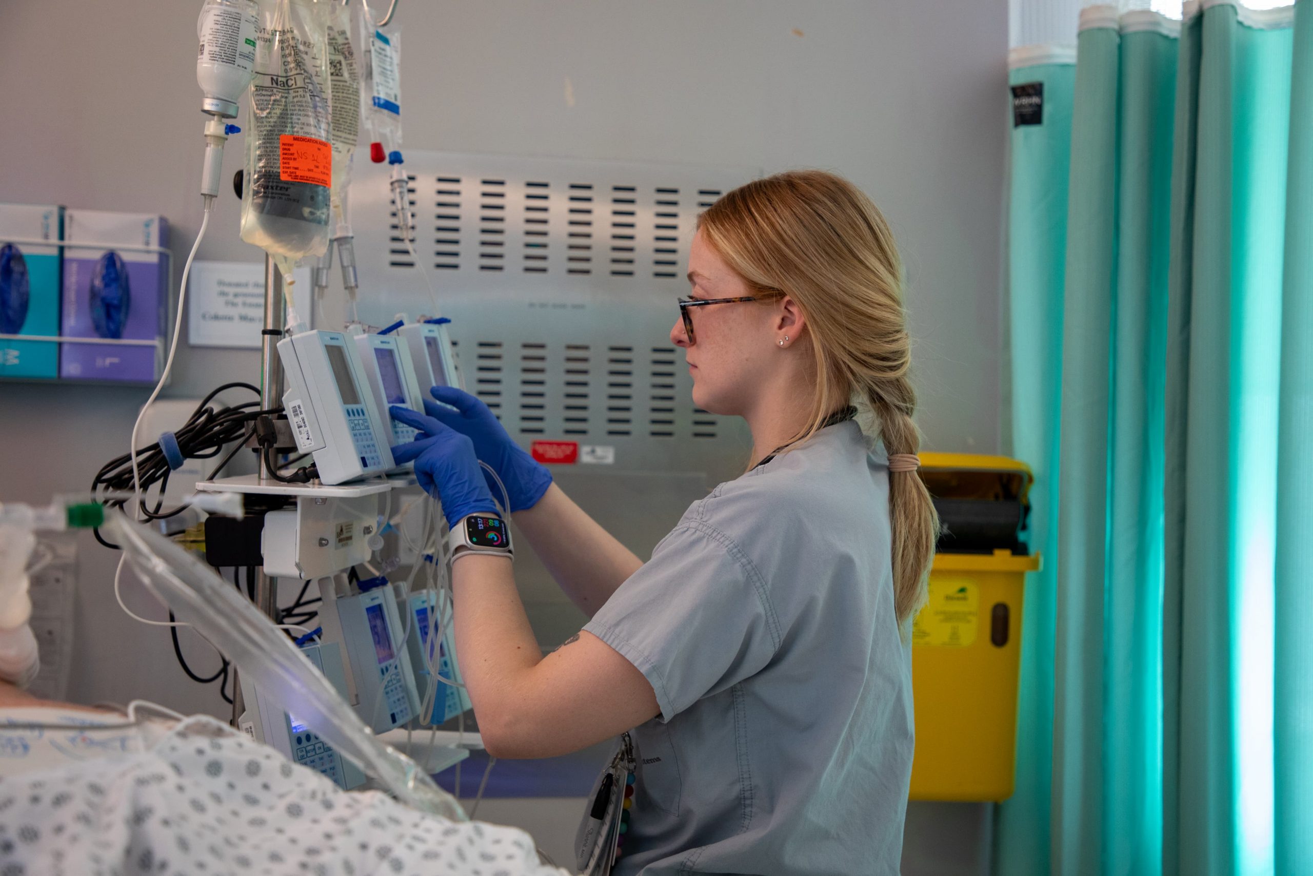A healthcare worker adjusts medical equipment in a hospital room next to a patient’s bed.