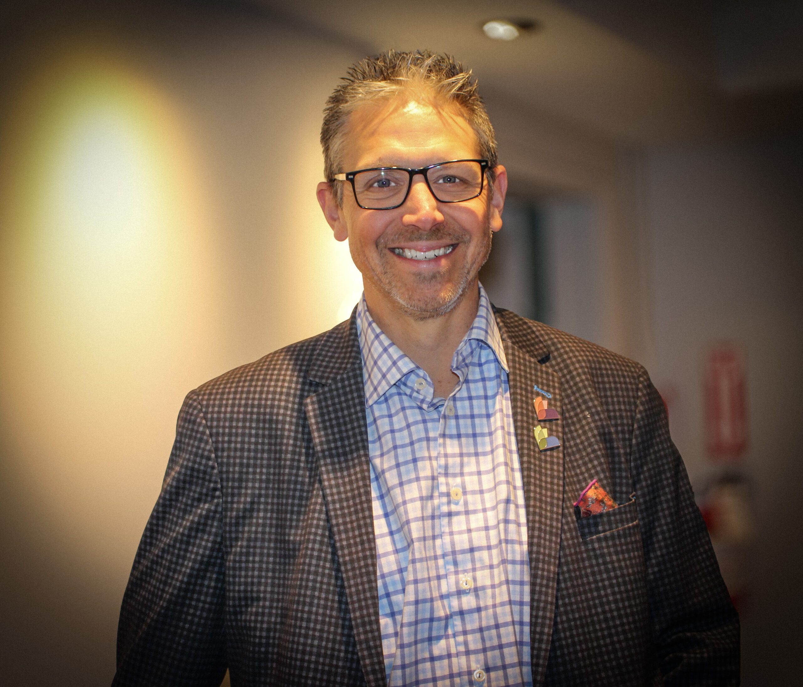 Tim wearing glasses, a checked suit jacket, and a plaid shirt, smiling indoors with soft lighting.
