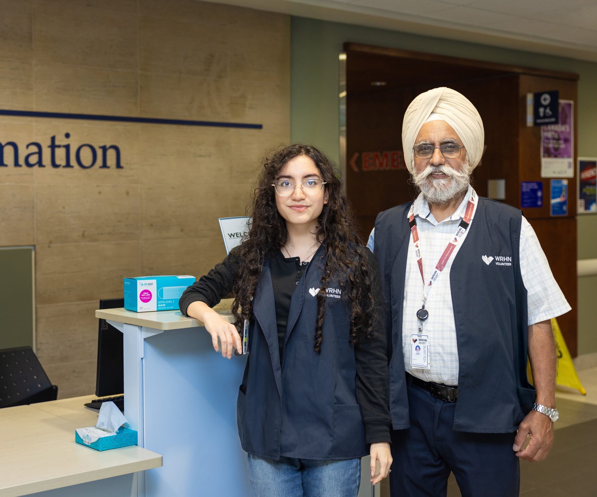 Two staff members stand at an information desk in a public building, both wearing matching vests and name badges.