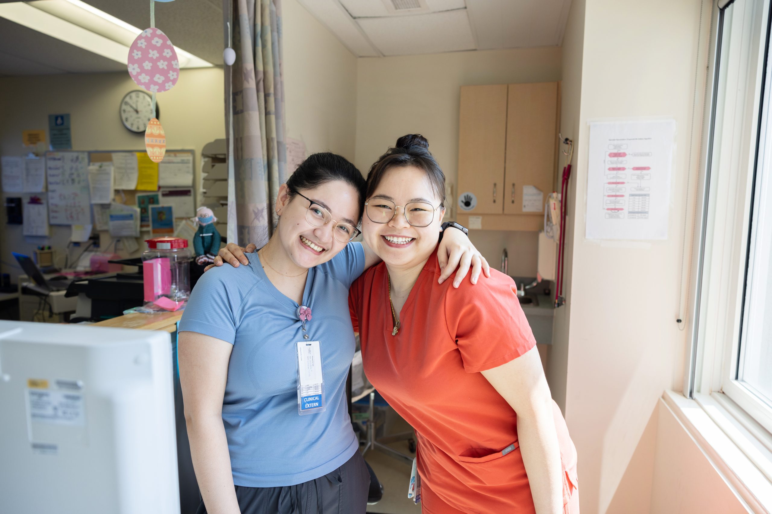 Two healthcare workers in scrubs stand smiling with arms around each other in a brightly lit medical office.